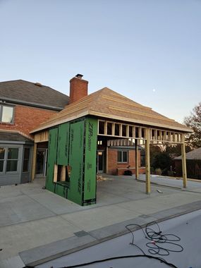 Construction of a patio cover attached to a brick house with exposed framing, green sheathing, and a pool in the foreground.
