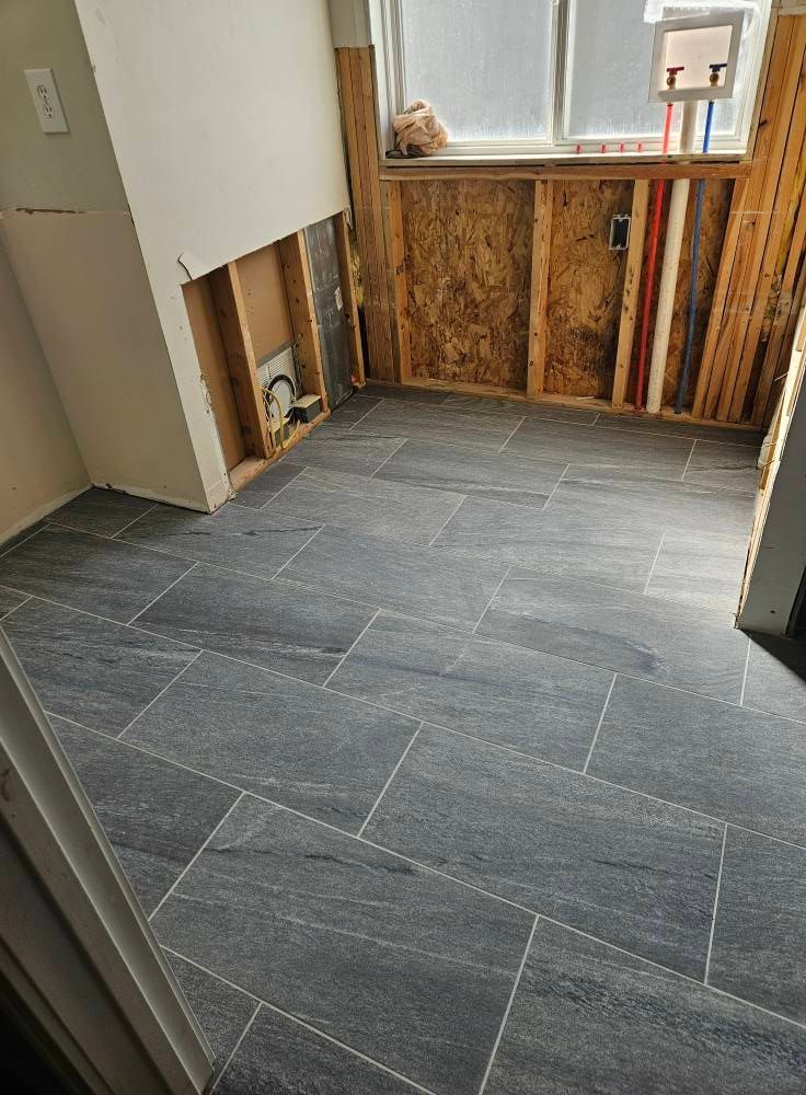 Gray tile floor in a room with unfinished walls and exposed pipes near a window.