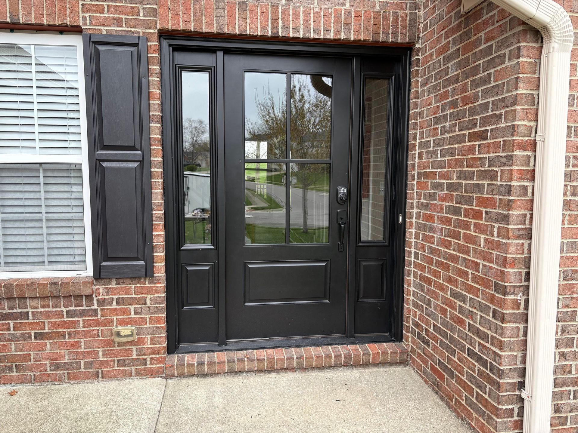 Black front door with sidelights and shutters on a brick house.