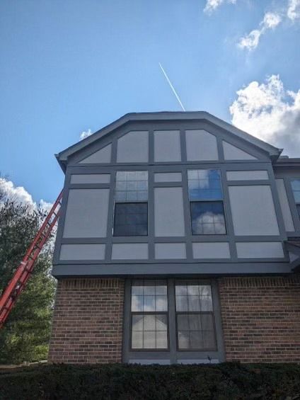 House with gray and white paint, brick base, ladder, blue sky with contrail.