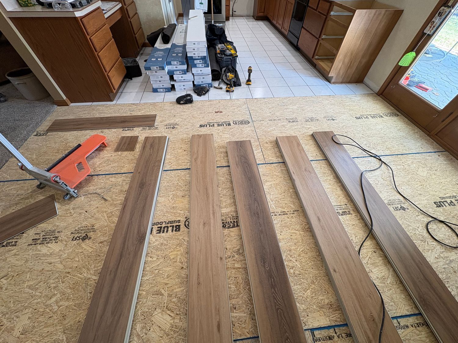 Flooring installation in progress. Wood planks laid out on subfloor with tools and cabinets in the background.