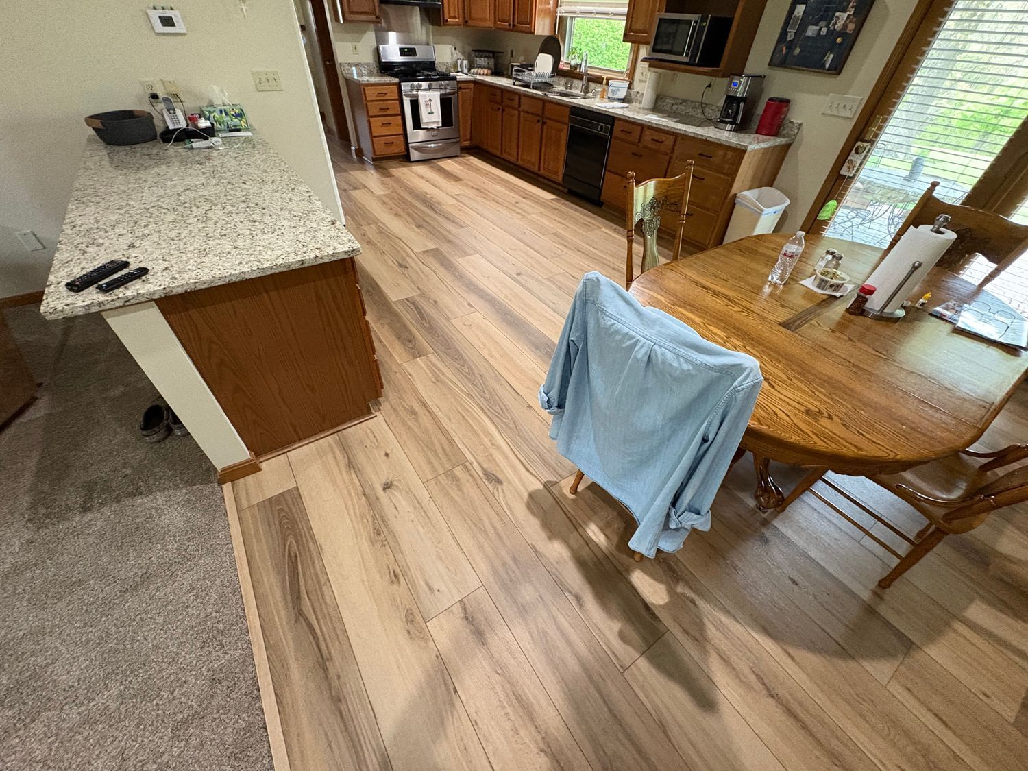 Kitchen with wooden floor, cabinets, and a granite countertop. A blue shirt hangs on a chair by a round wooden table.