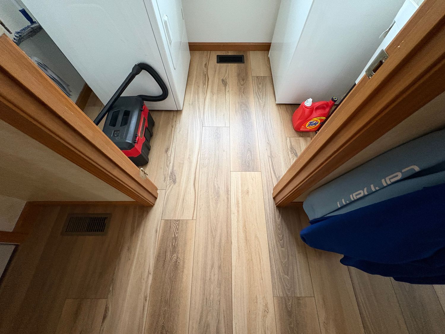 Narrow hallway with wood-look flooring, flanked by white cabinets, a red vacuum, and a blue cushion.