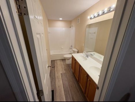 Bathroom with double sink vanity, toilet, and bathtub. Gray wood-look floor and beige walls.