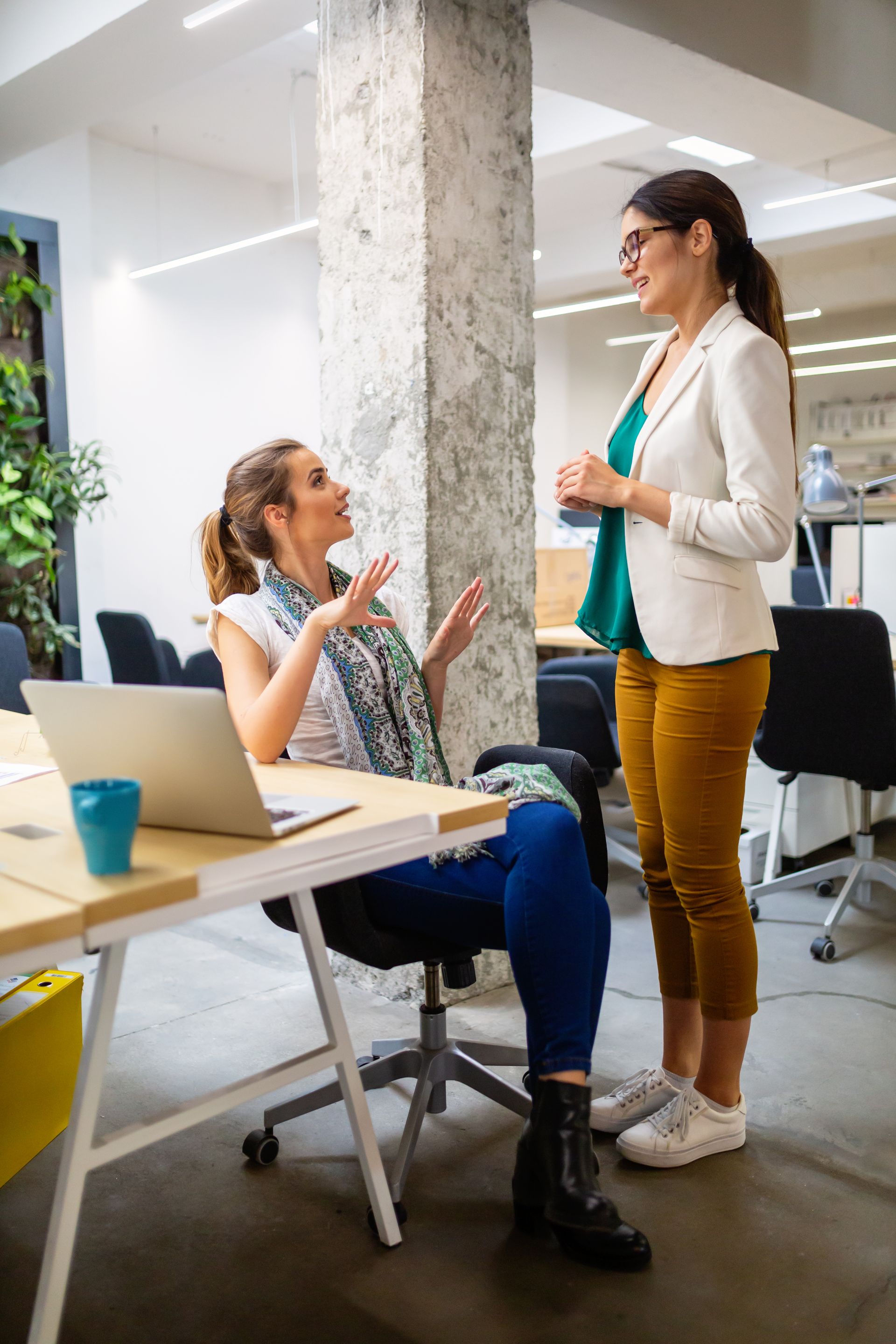 Two women are having a conversation in an office while one woman is sitting at a desk with a laptop.