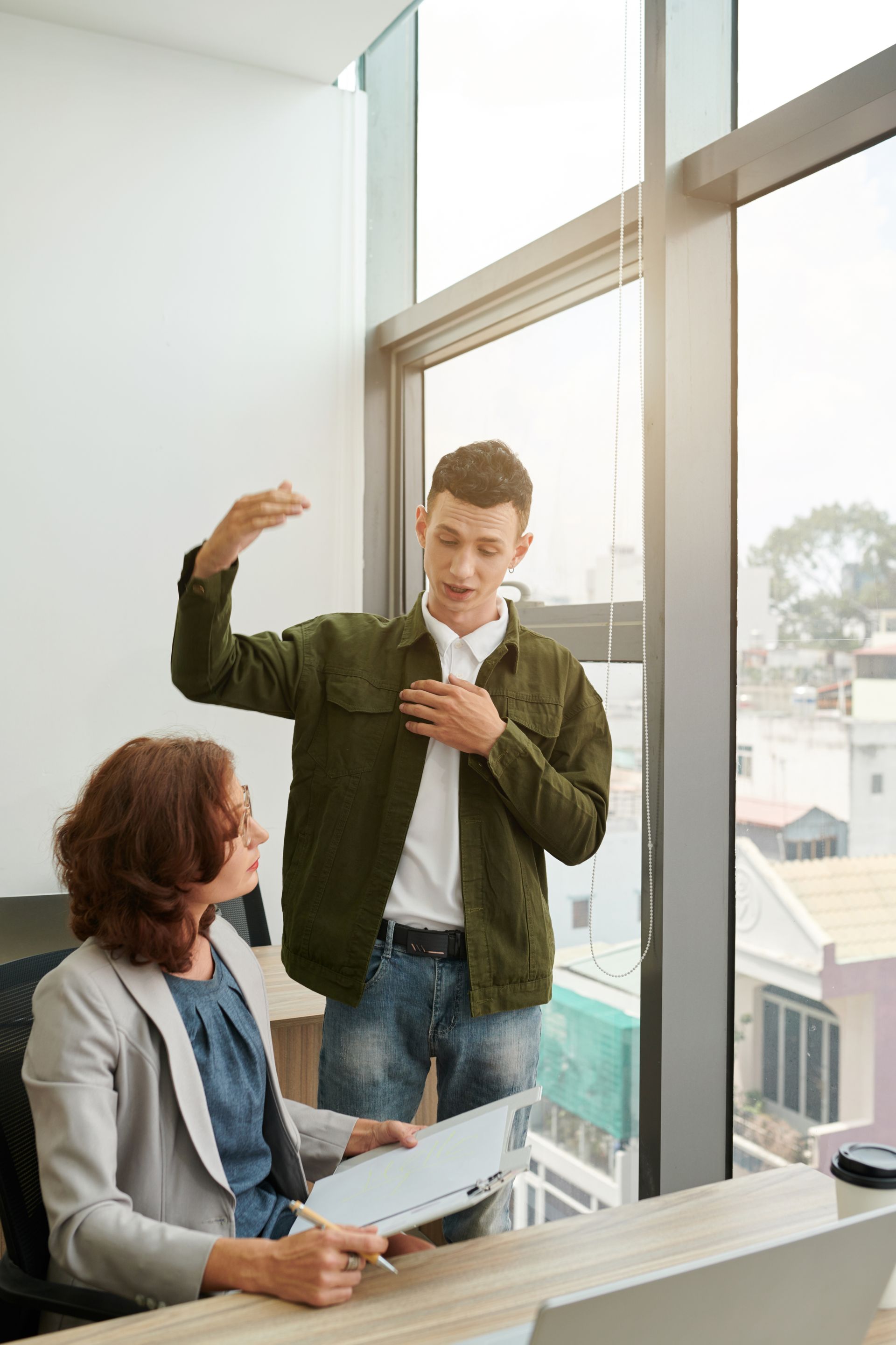 A man is standing next to a woman sitting at a desk in front of a window.