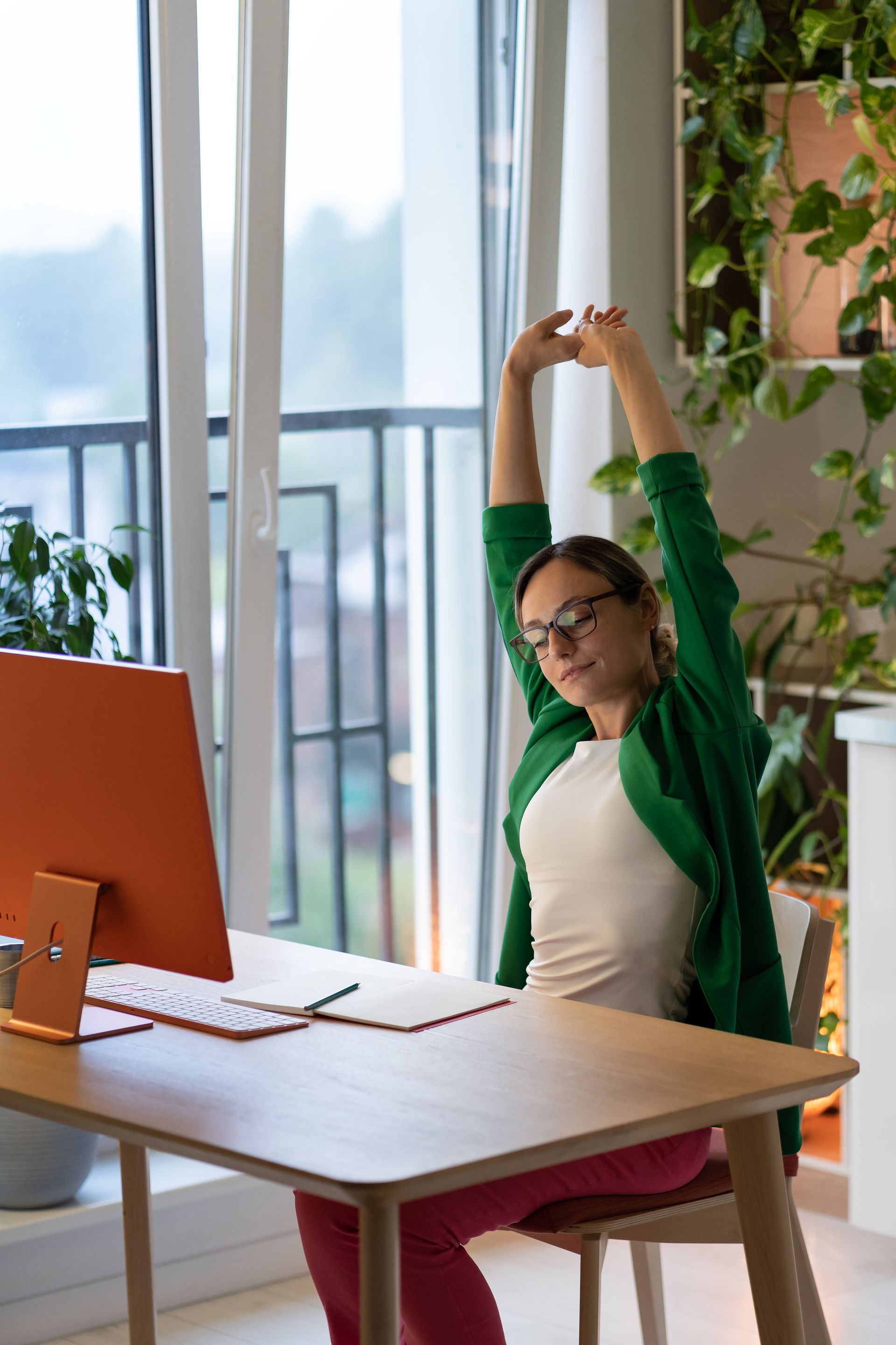 A woman is stretching her arms while sitting at a desk in front of a computer.