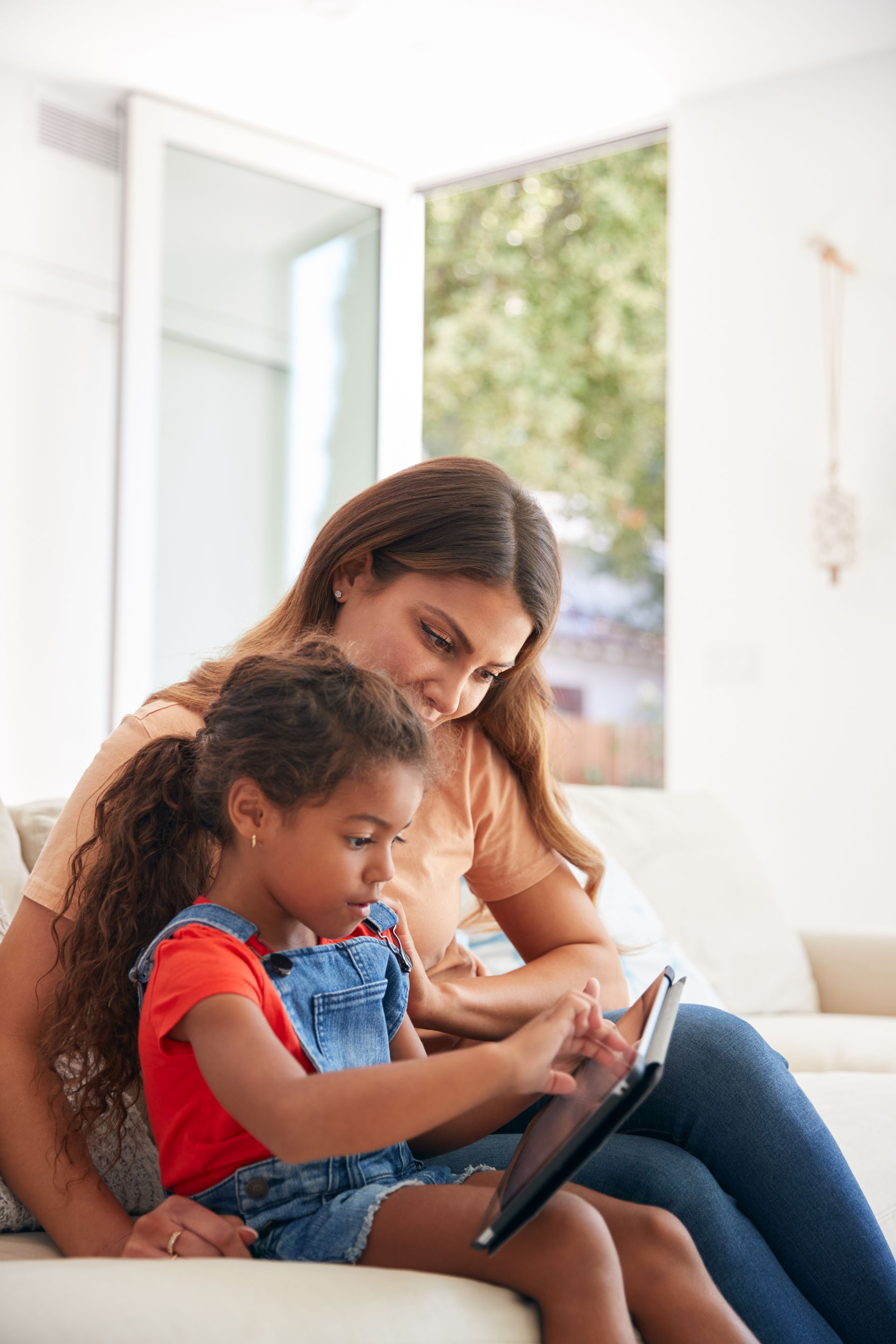 A woman and a little girl are sitting on a couch looking at a tablet.