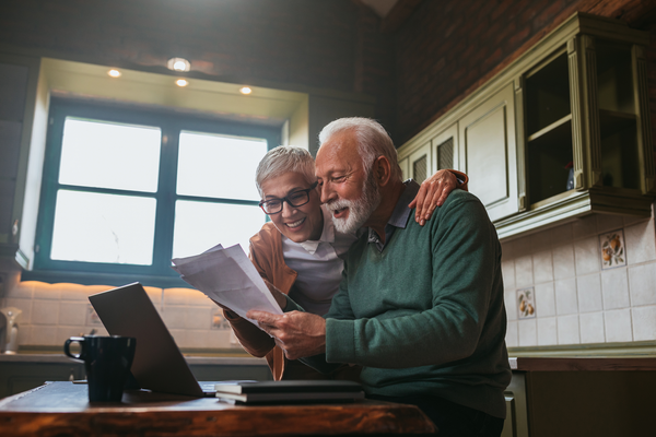 Two older men are sitting at a table in a kitchen looking at papers and a laptop.