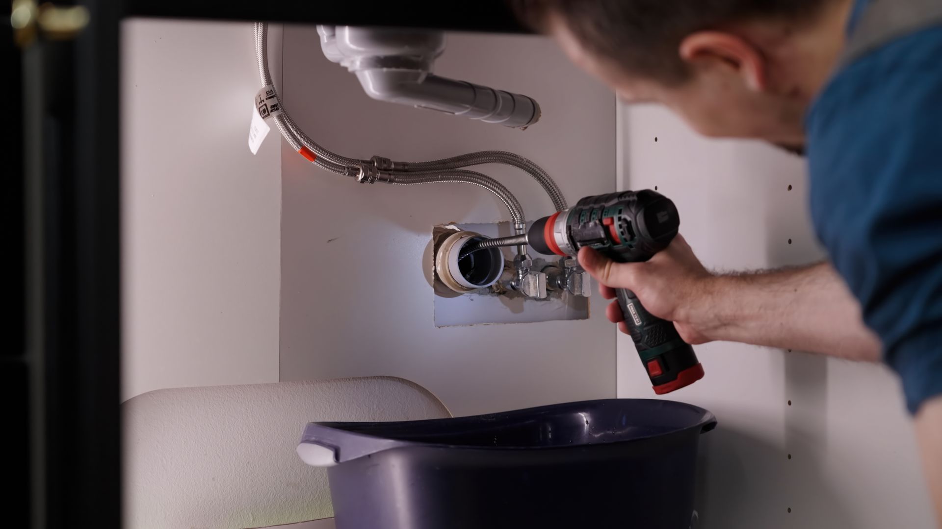 Man using a drill under a sink with a bucket to catch water Man using a drill under a sink with a bucket to catch water