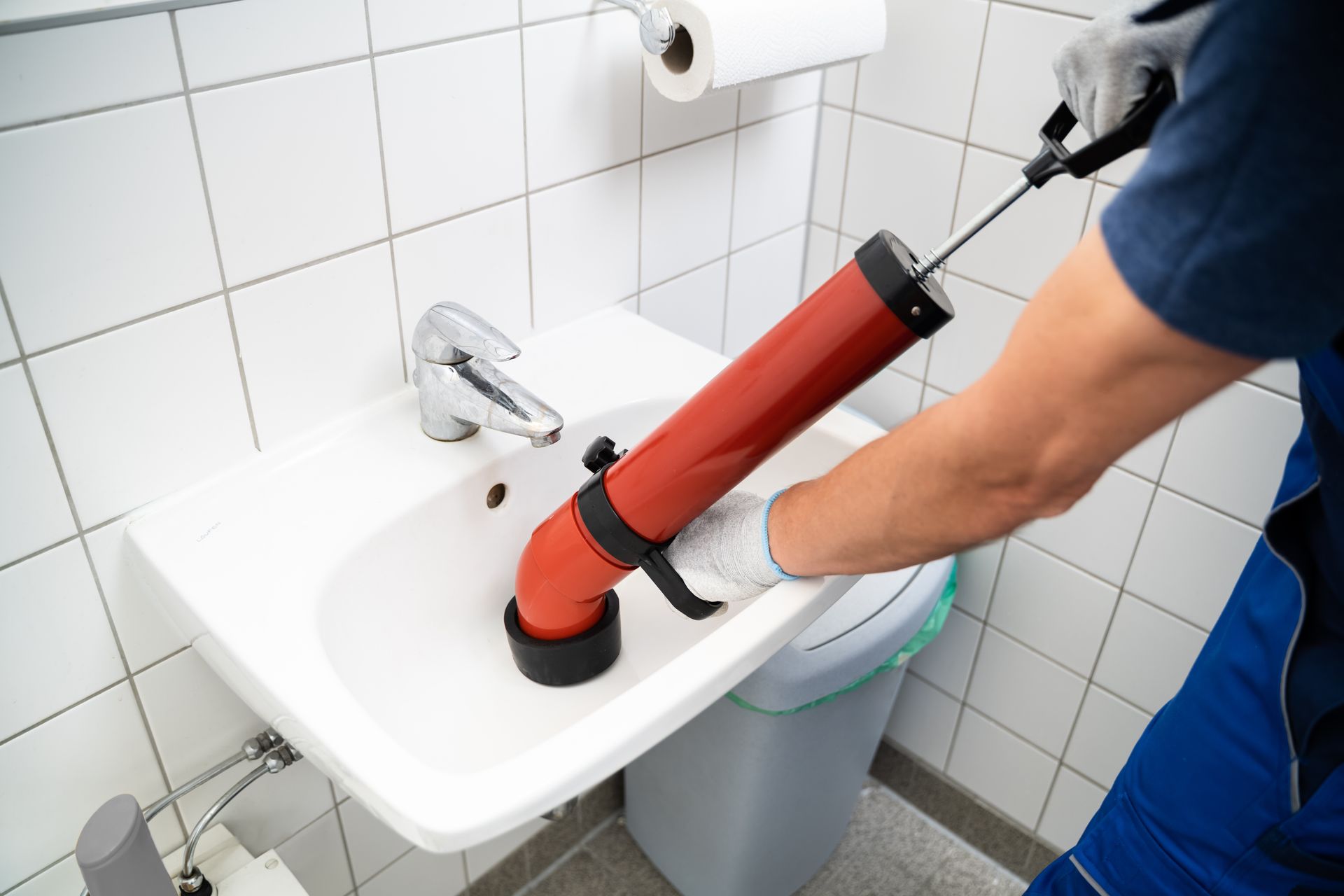 Plumber using a plunger to unclog a white sink in a tiled bathroom Plumber using a plunger to unclog a white sink in a tiled bathroom