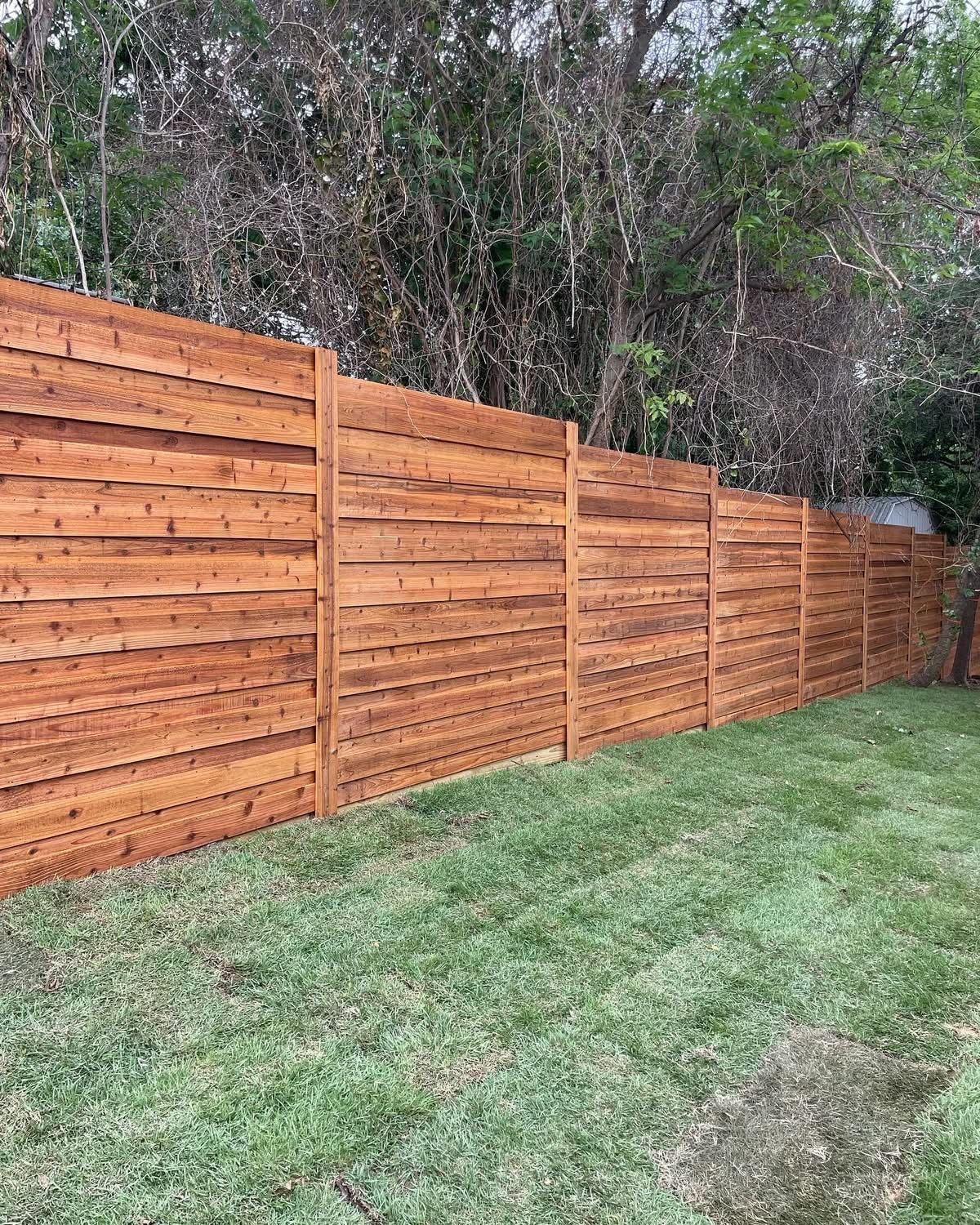 Wooden privacy fence stained brown in a grassy backyard, trees in the background.