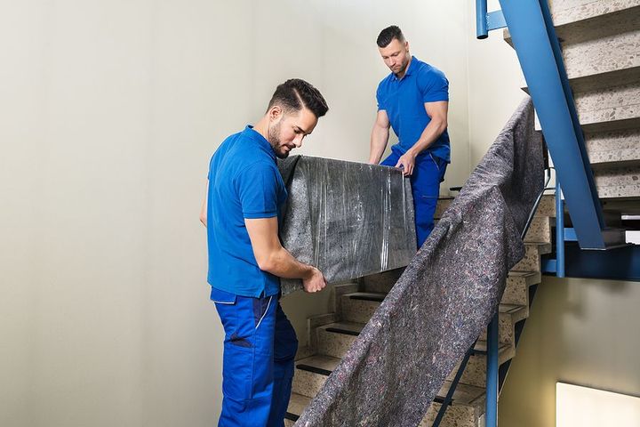 Two movers in blue uniforms carrying a heavy wrapped box up a staircase.