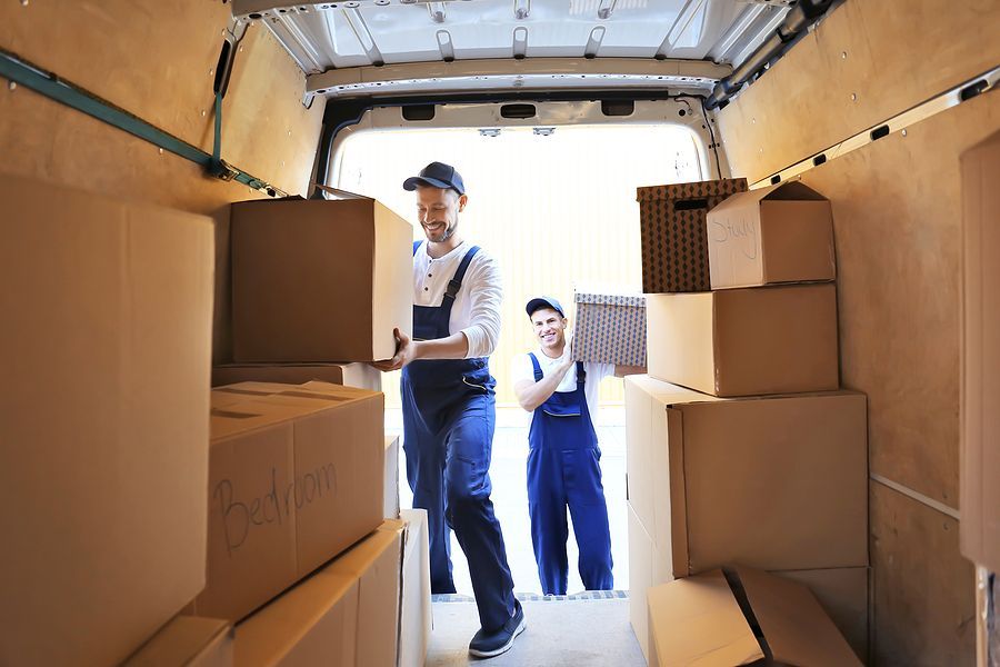 Two smiling movers in blue overalls loading cardboard boxes into a moving van.