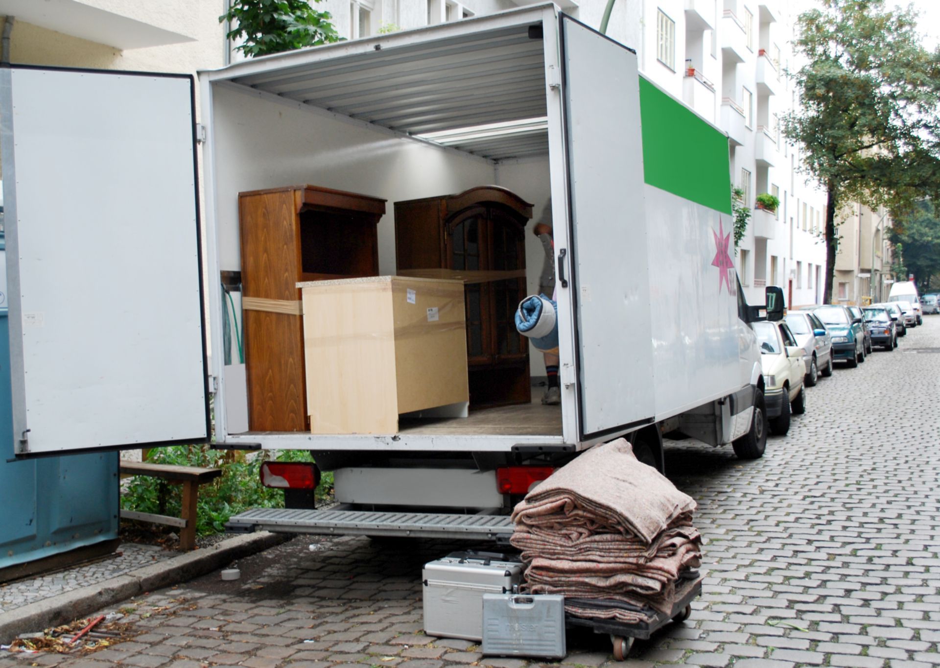 Open moving truck on a cobblestone street loaded with wooden furniture and moving blankets.
