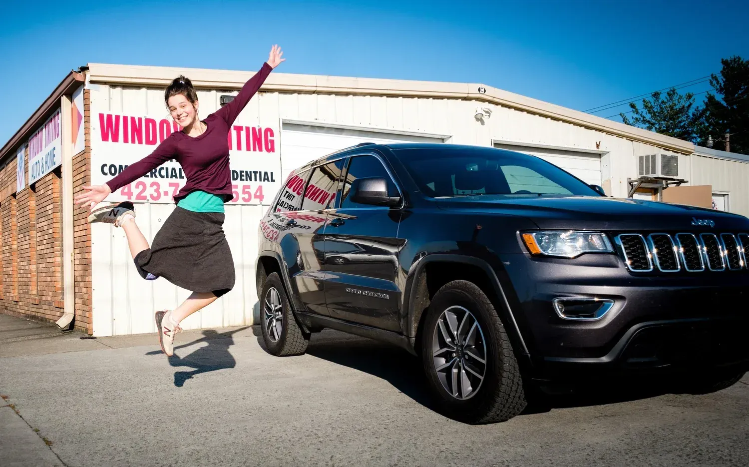 Woman jumps for joy near a dark SUV in front of a building with window tinting signage.