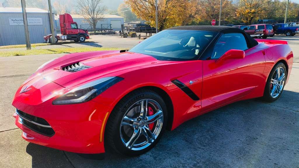 Red convertible Corvette parked on pavement with a semi-truck in the background.