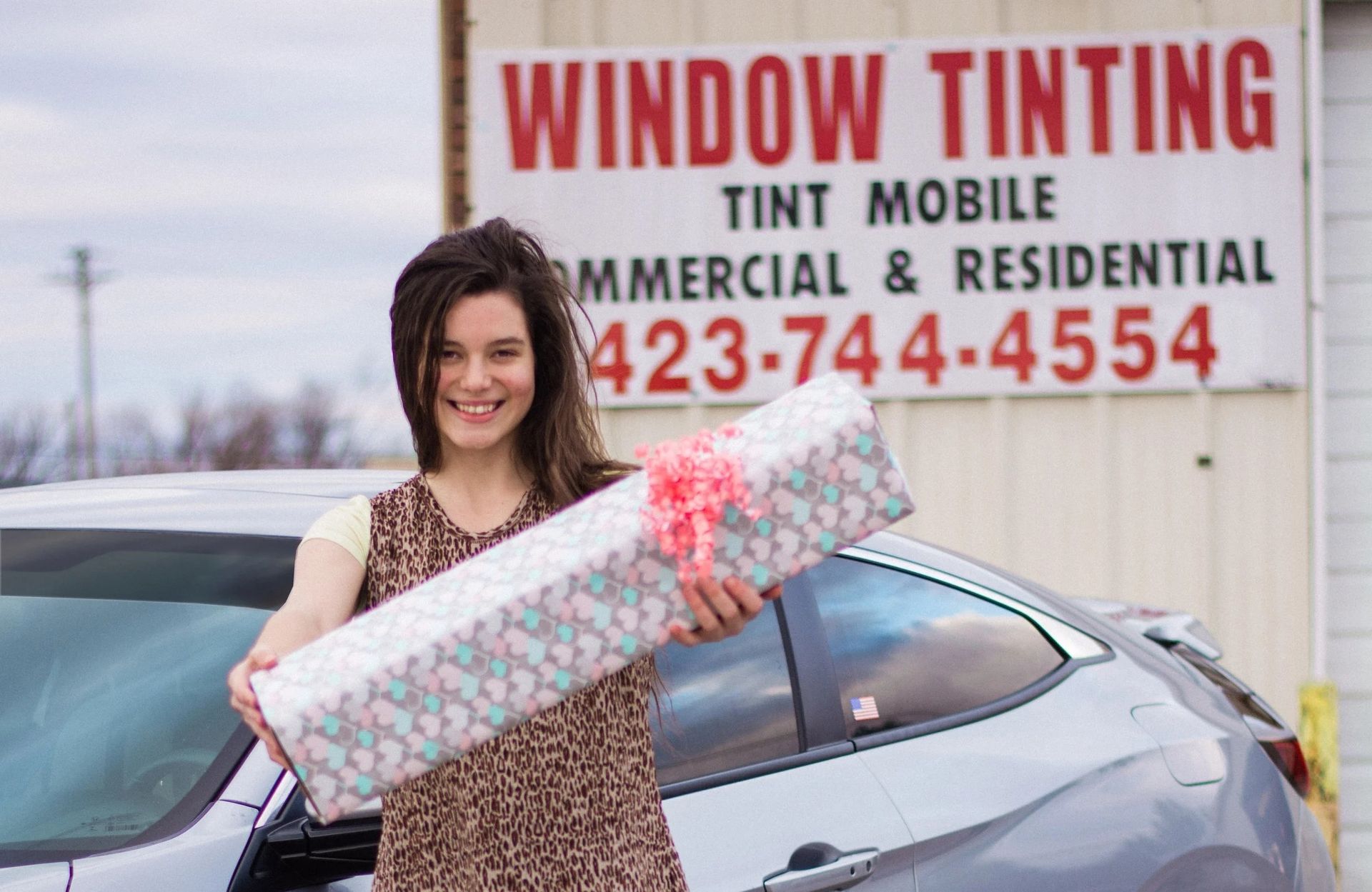 Woman holding wrapped gift in front of a car; window tinting sign in background.