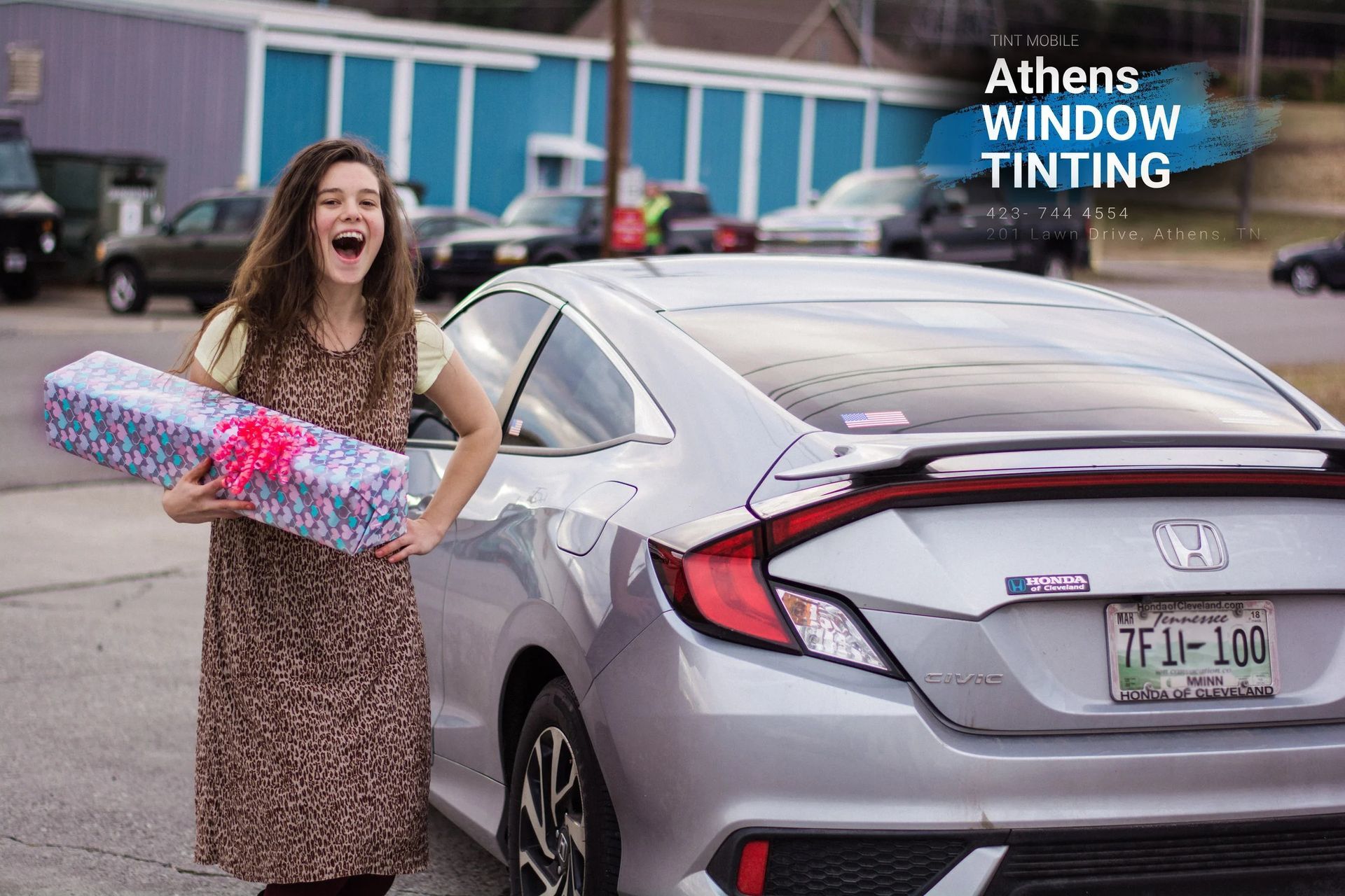 Woman holding a gift beside a silver car, joyful expression. Athens Window Tinting logo in corner.