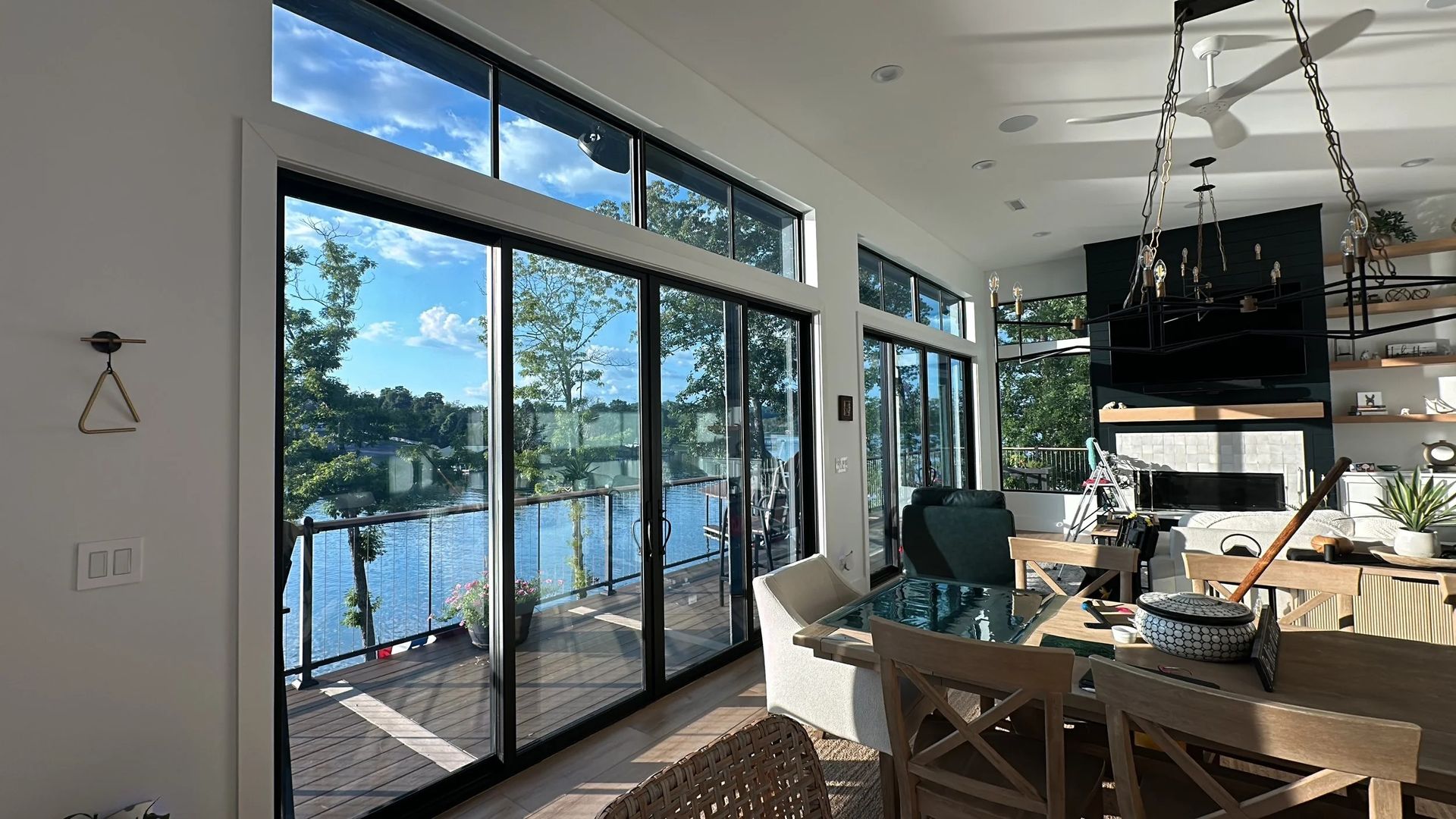 Bright interior with large windows overlooking a lake. A dining table and fireplace are visible.