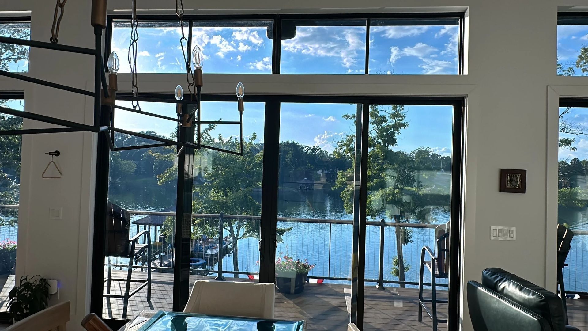 Interior view through large windows overlooking a lake. A chandelier hangs; light blue sky and trees are visible.
