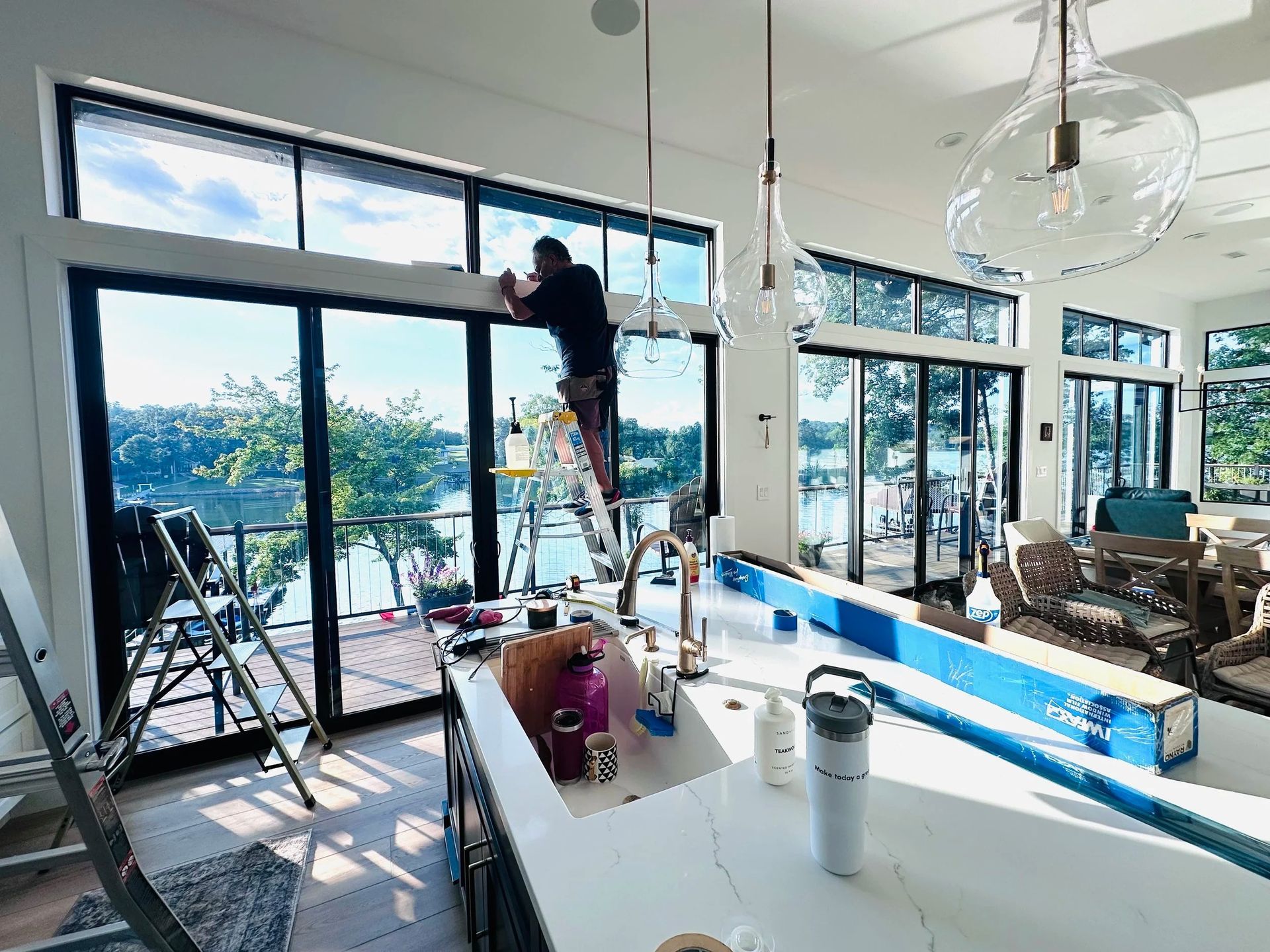 Man on a ladder installing a window treatment over large windows overlooking a lake.