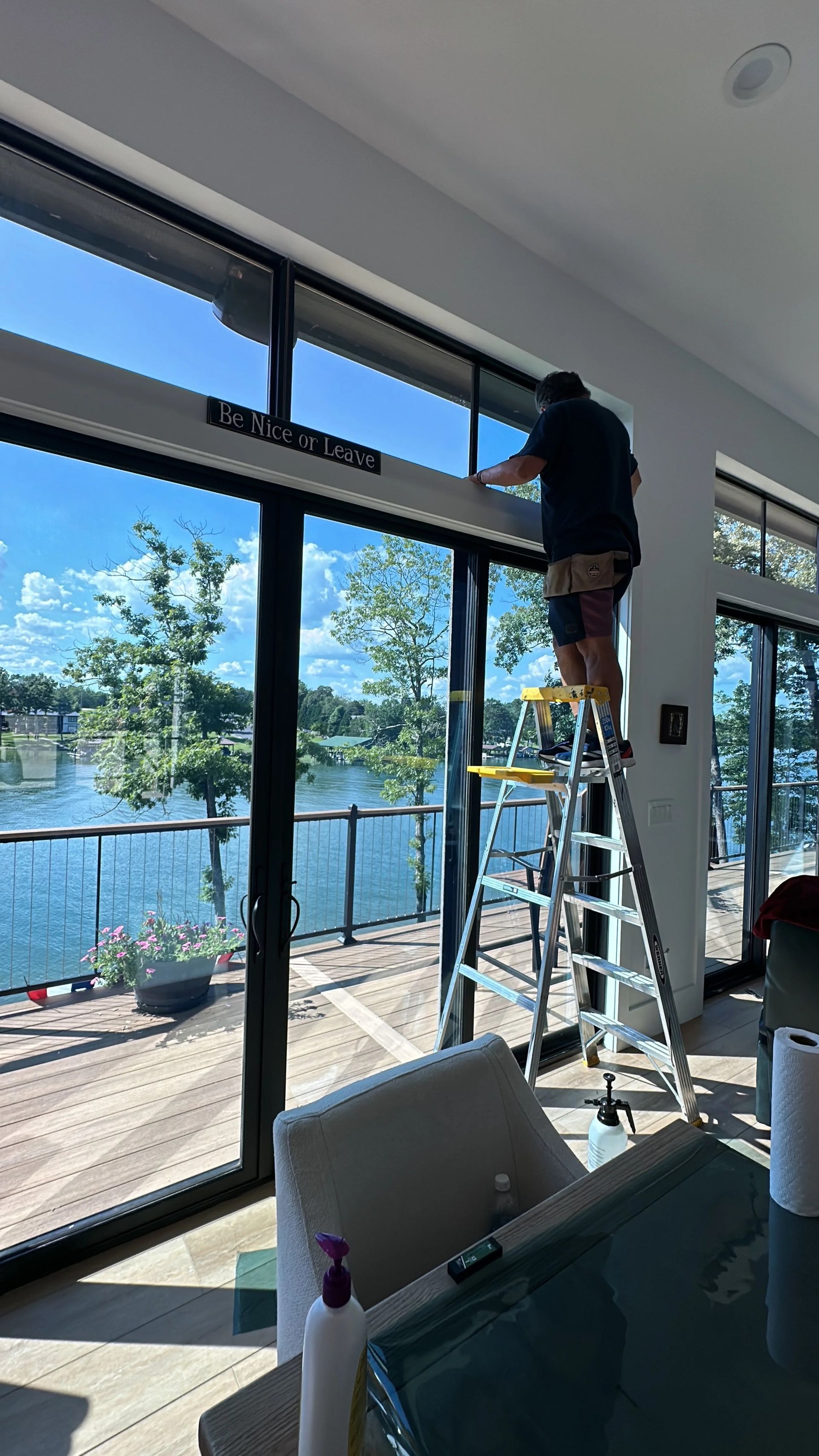 Person on a ladder installing window film on large glass doors overlooking a lake.