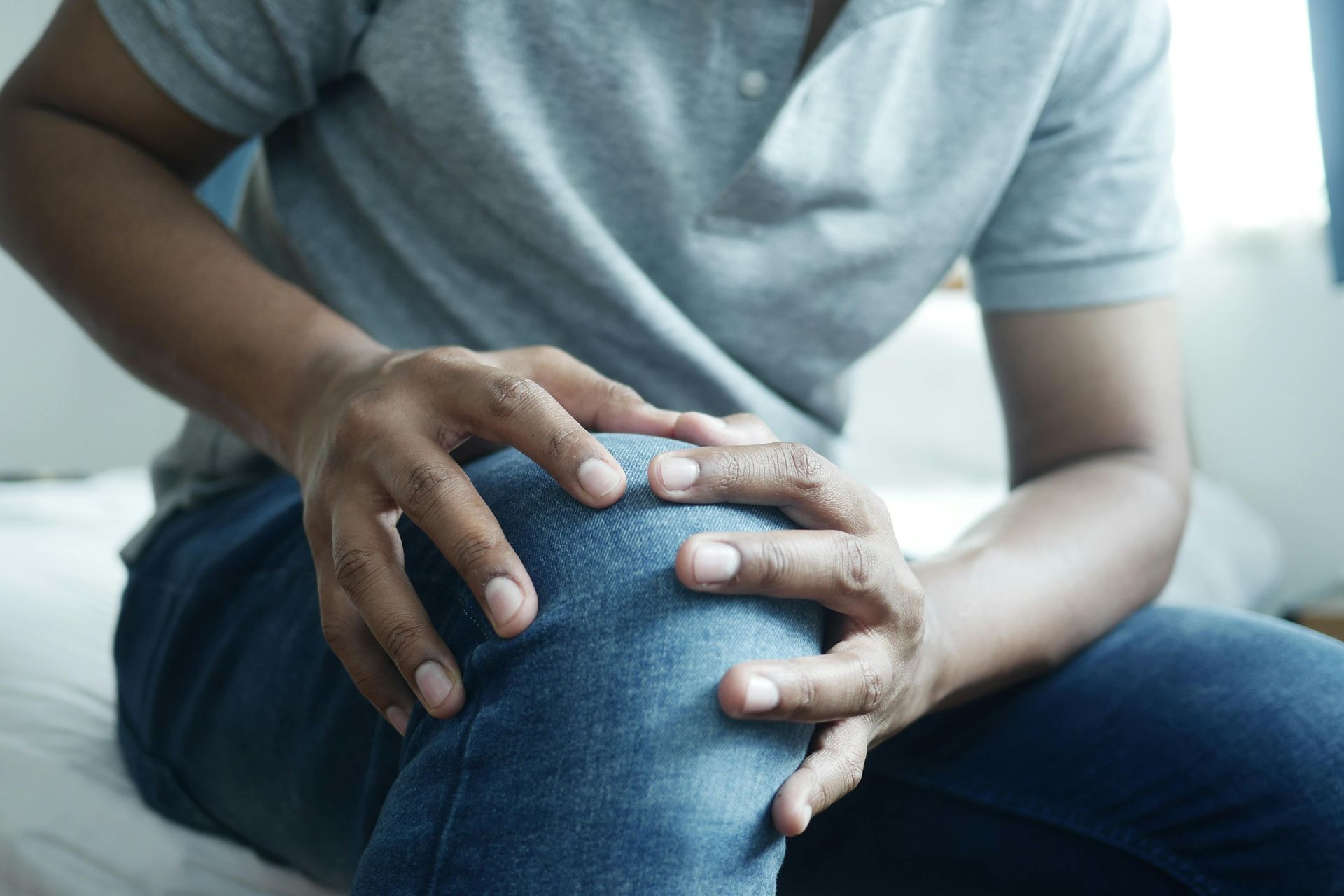 A person in a gray shirt and blue jeans sitting on a bed while holding their knee with both hands in visible discomfort.