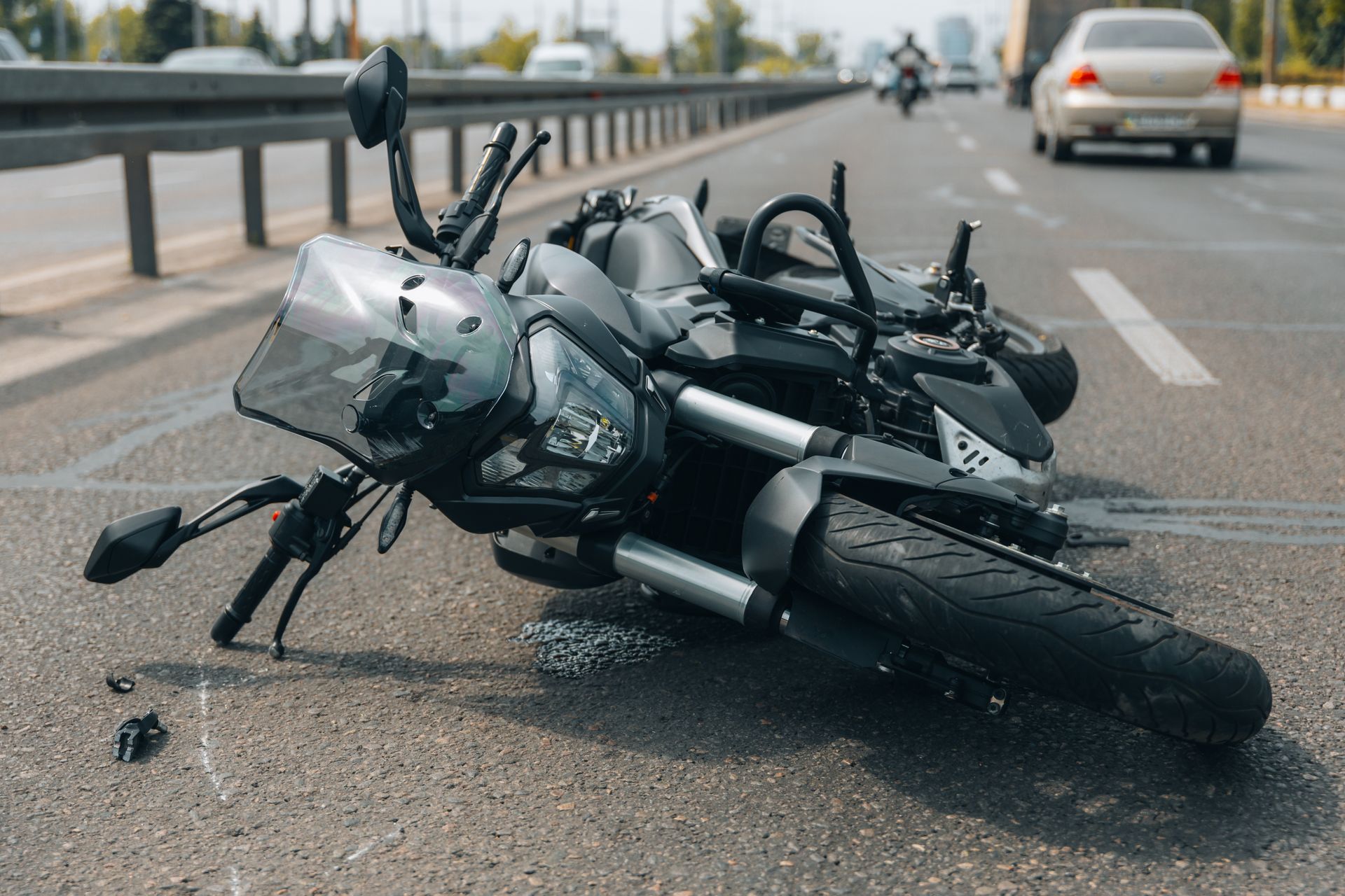 A black motorcycle lies on its side on an asphalt highway with a blurred car and another motorcycle in the background.
