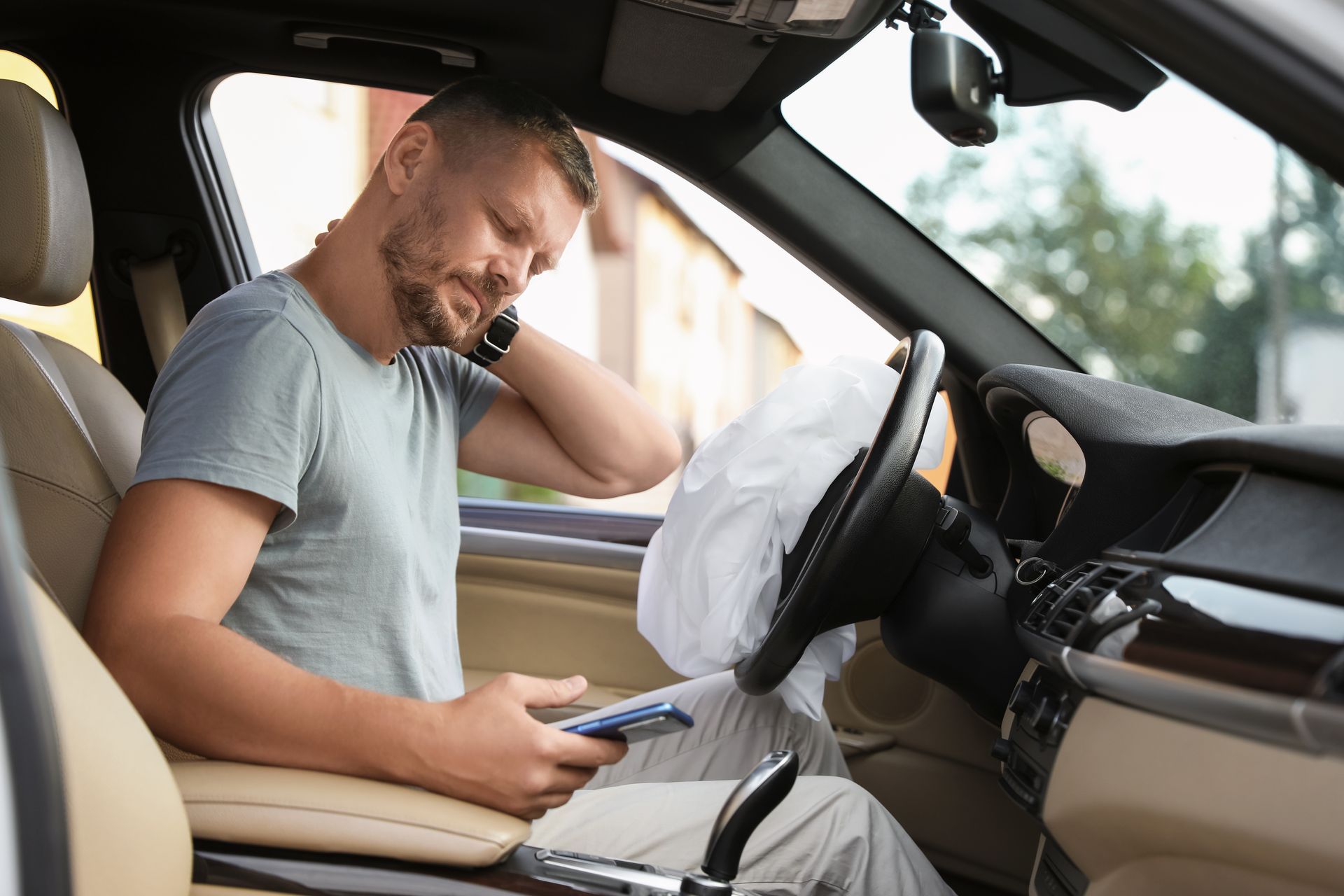 A person sits in a car with a deployed airbag, holding a phone and clutching their neck with a pained expression.