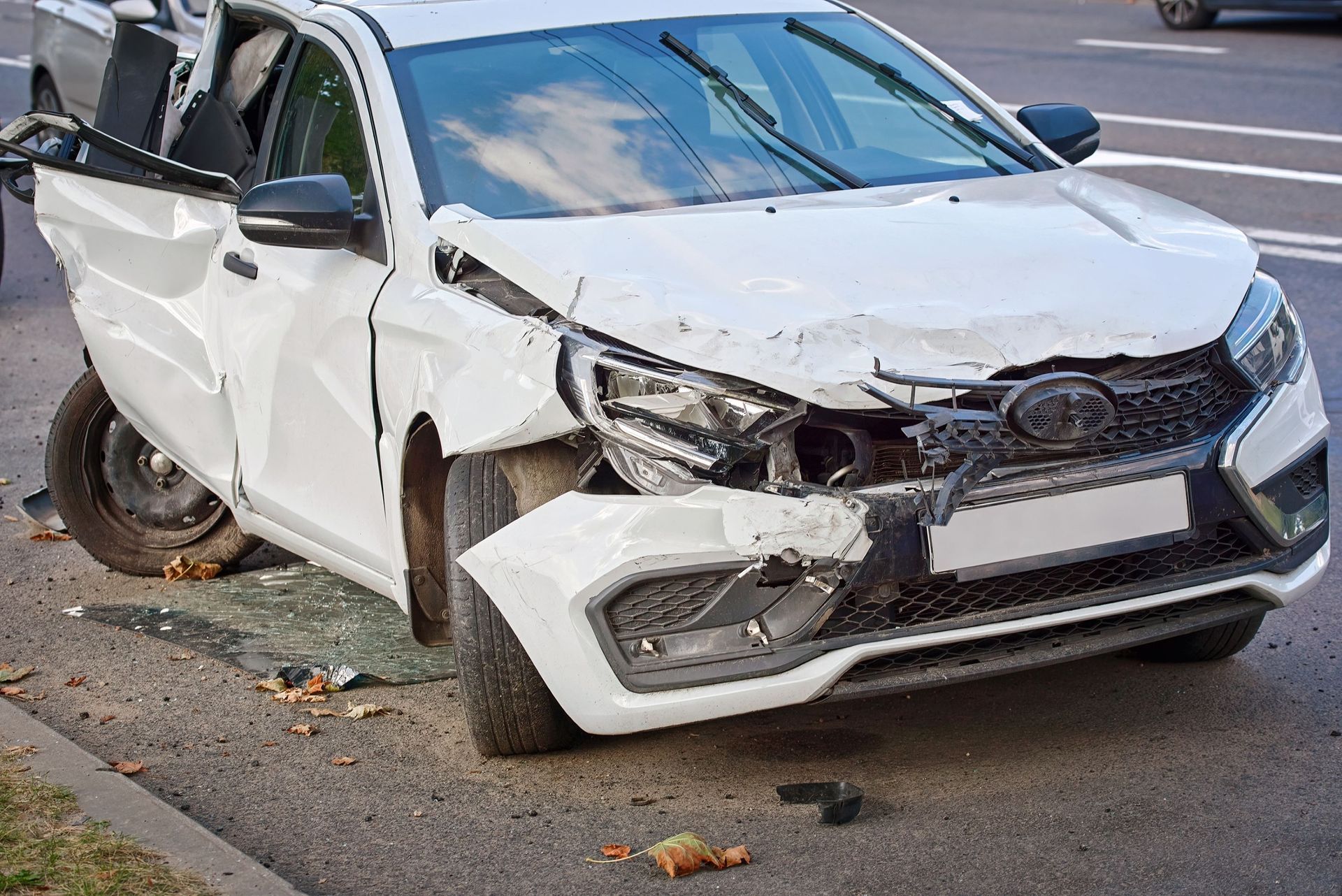 A damaged white car on the side of a road with a crushed front bumper and a shattered side window.