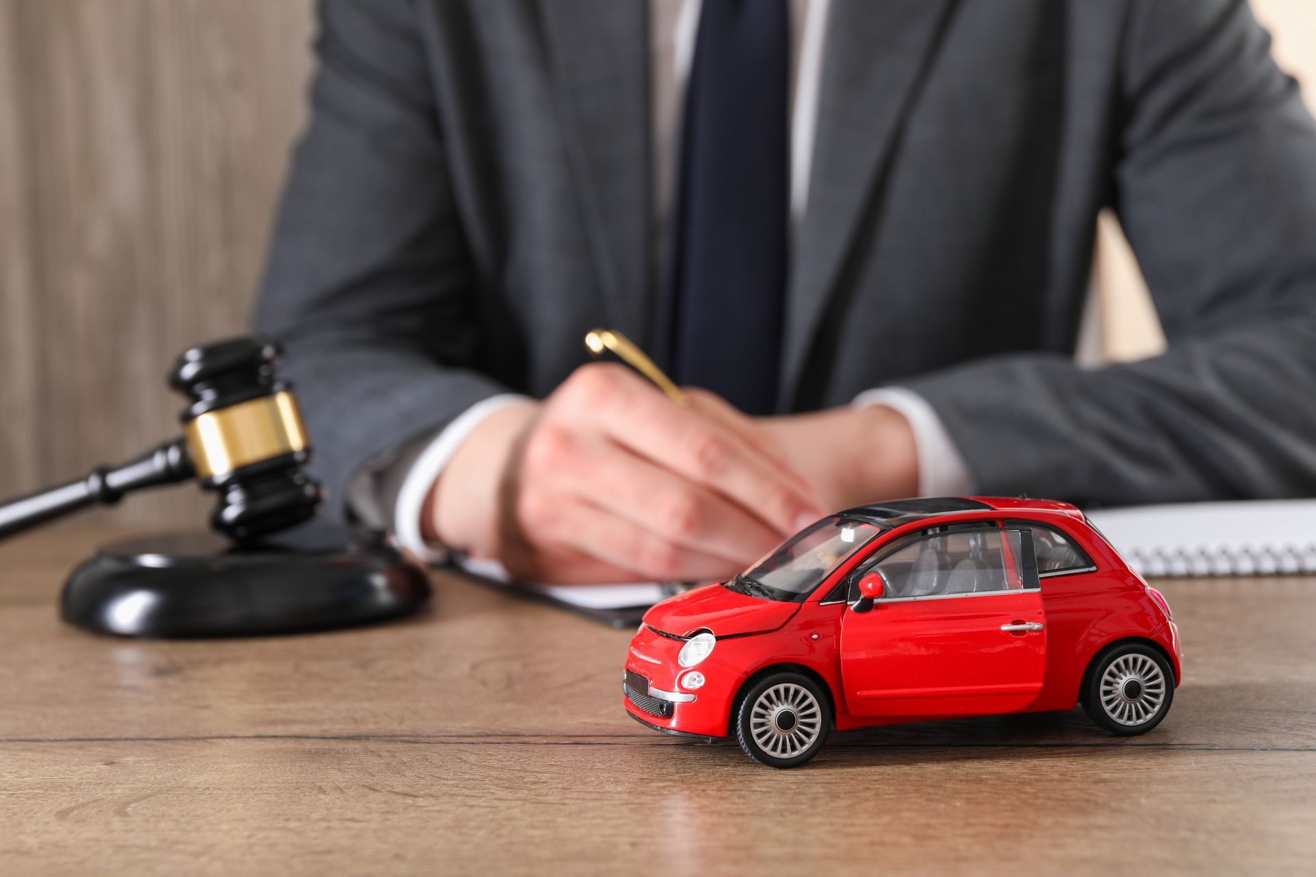 A person in a suit sits at a desk with a gavel and a small red toy car model.