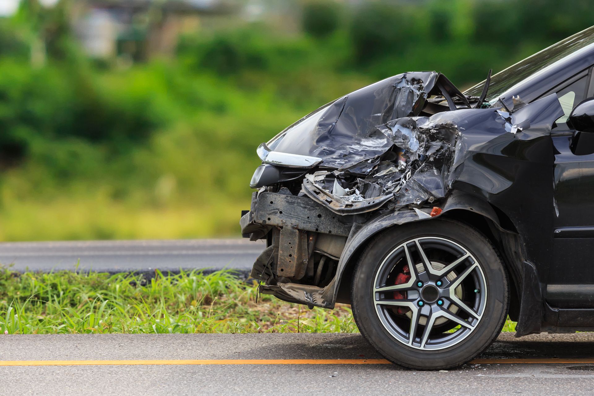 A heavily damaged black car sits on the shoulder of a road with its front hood crumpled.