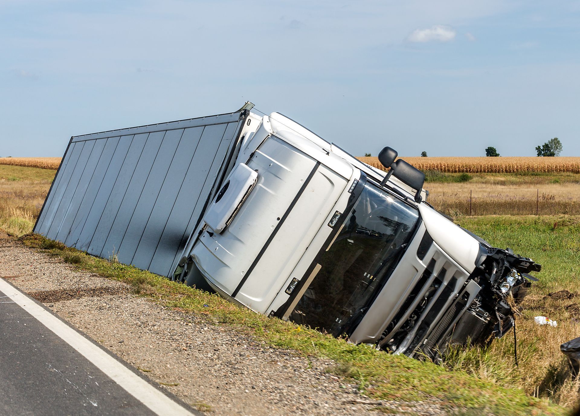 A white semi-truck lies on its side on the grassy embankment next to an asphalt road in a rural field.