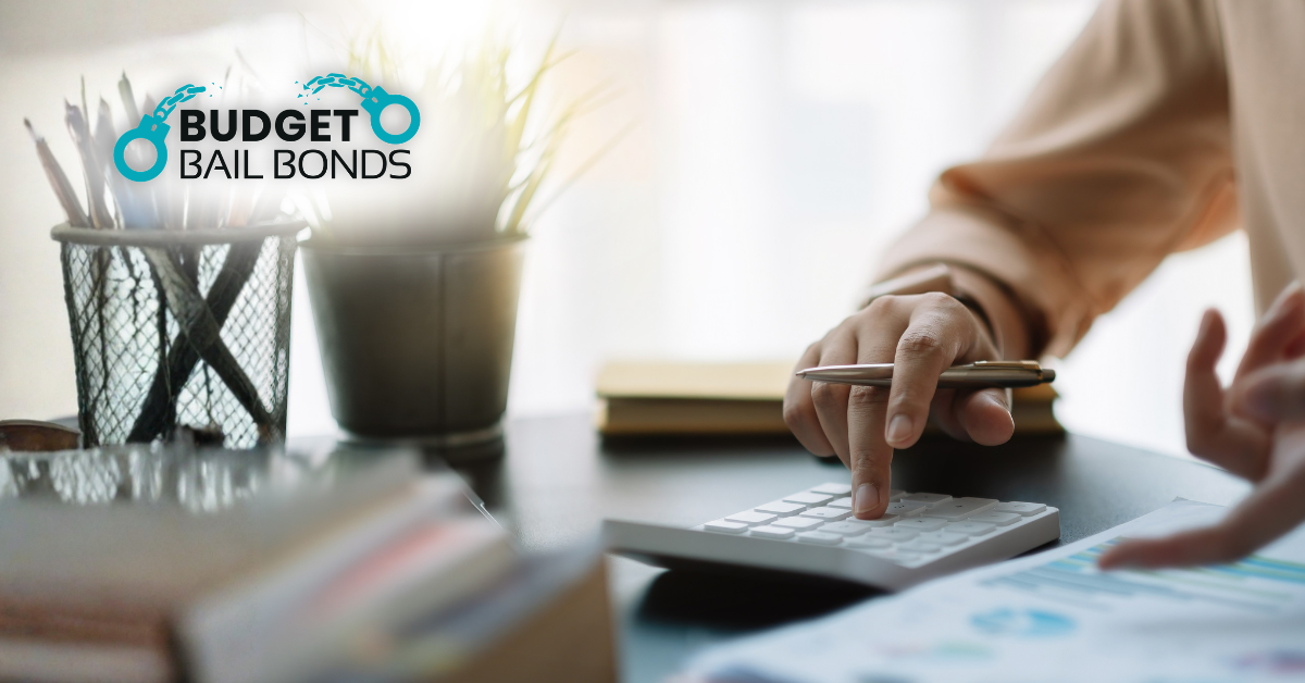 A person sits at a desk using a calculator, with a Budget Bail Bonds logo overlaid in the corner.
