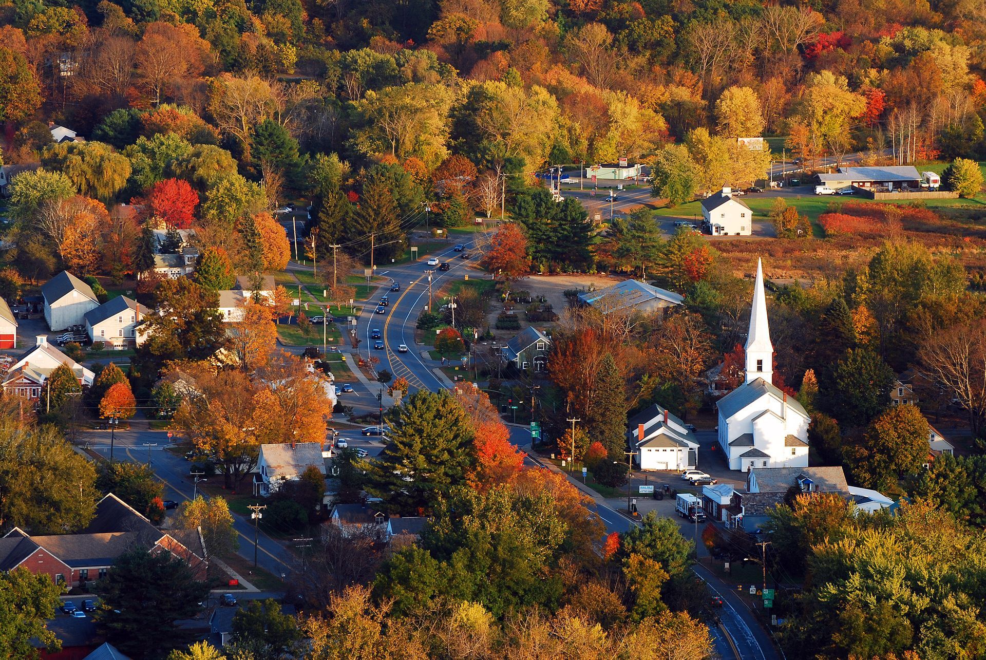 Aerial view of a town in autumn with colorful trees, a white church, and winding roads.