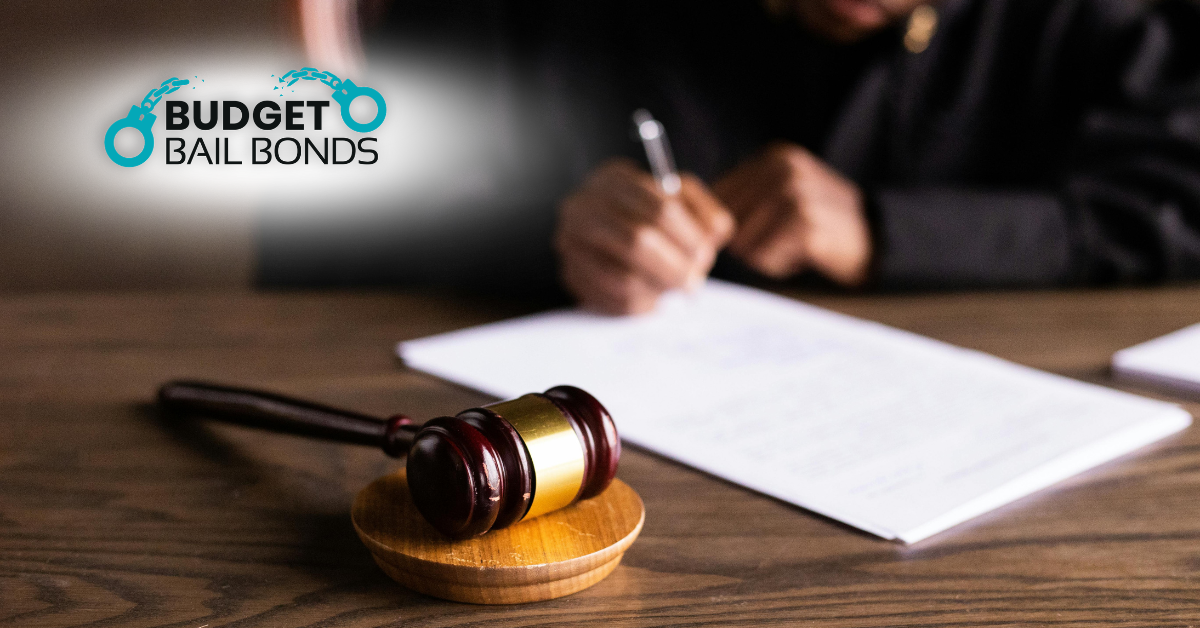 A wooden judge's gavel rests on a desk next to a person signing a document, with the Budget Bail Bonds logo above.