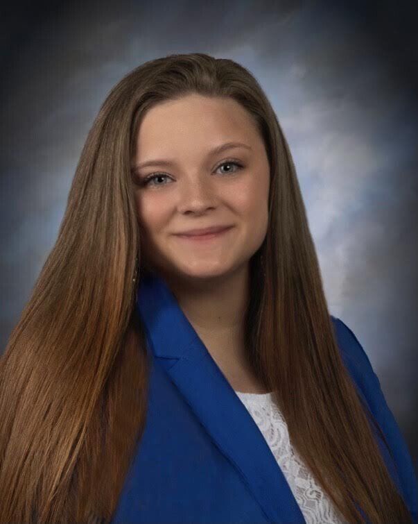 Woman with long brown hair, wearing a blue blazer and white shirt, smiling.