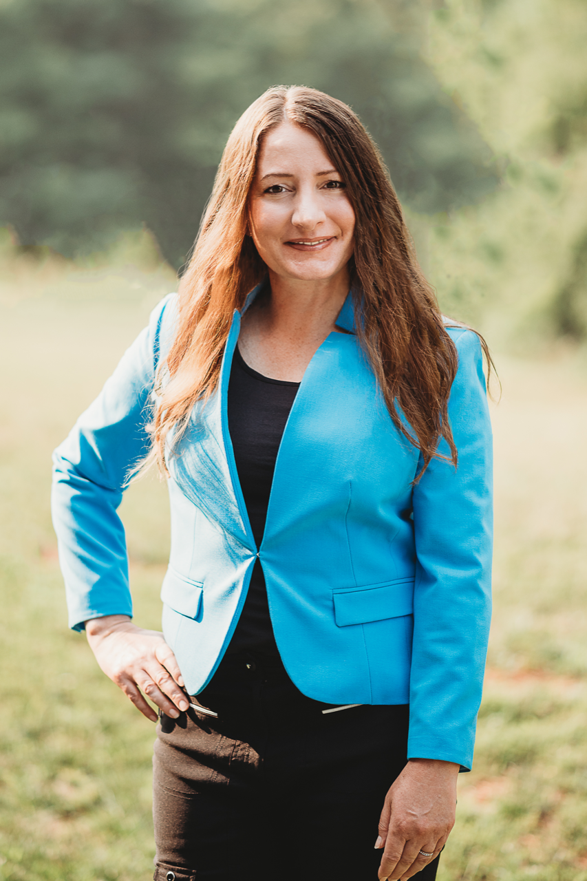 Woman with long brown hair in a blue blazer and black top, standing with hand on hip, outdoors.