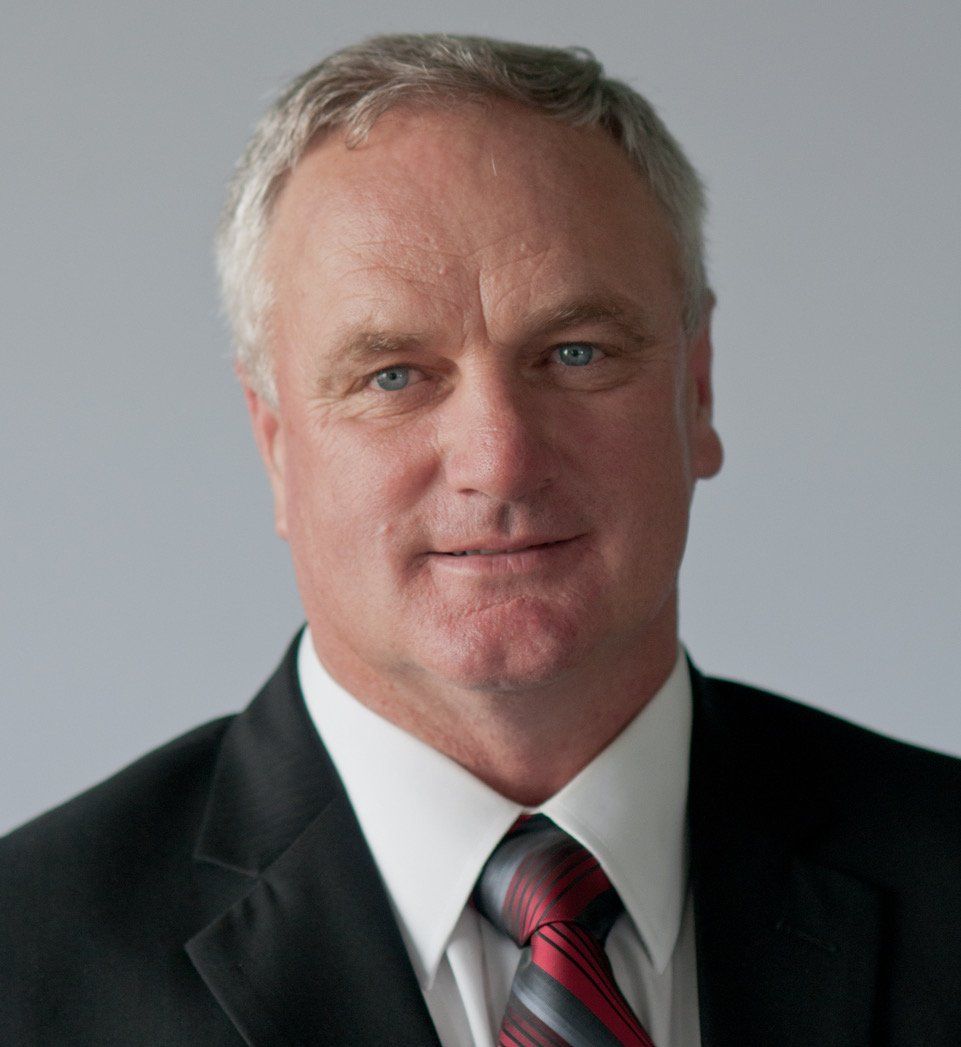Man in suit and tie, smiling, with gray hair, set against a gray background.