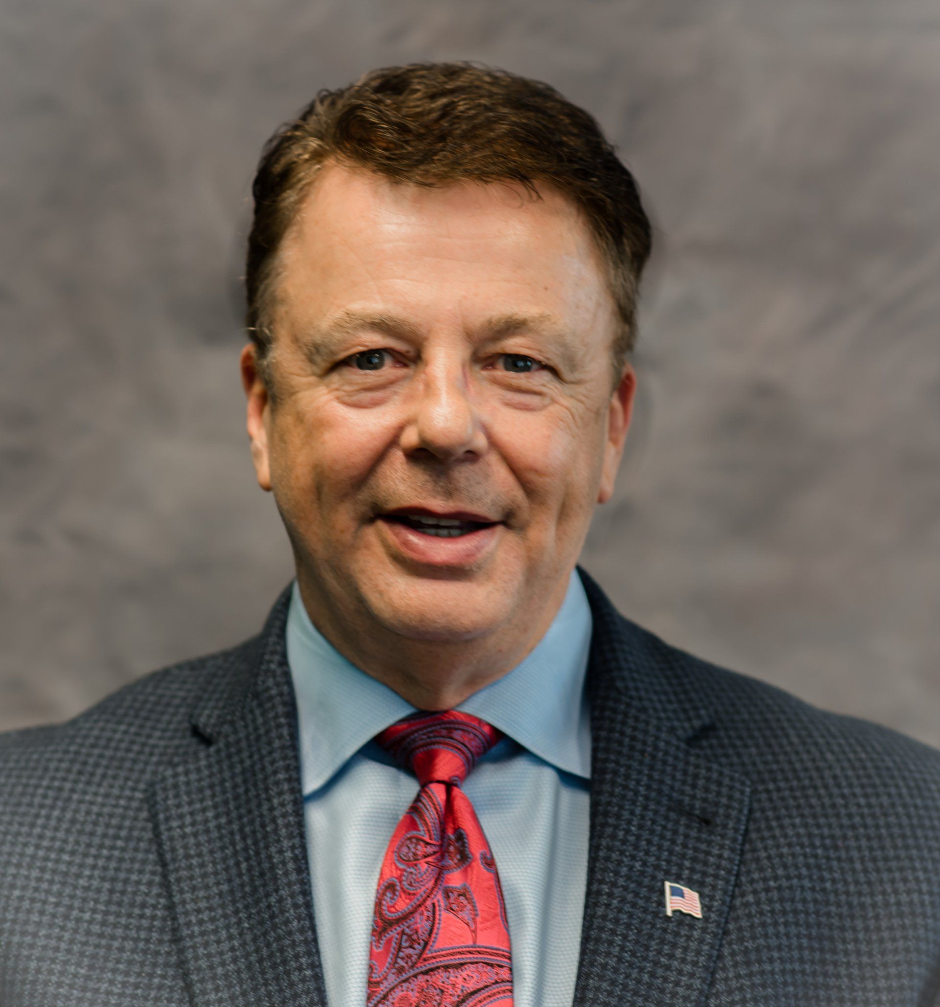 Man in suit, smiling, red tie, American flag pin.