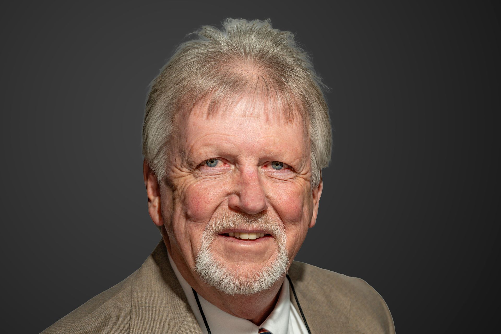 Man with gray hair and beard smiles, wearing a beige suit jacket against a dark gray background.