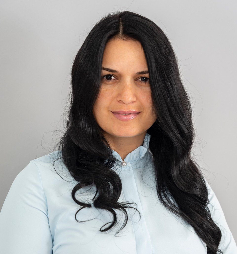 Woman with long black hair, smiling, wearing a light blue collared shirt, against a gray background.