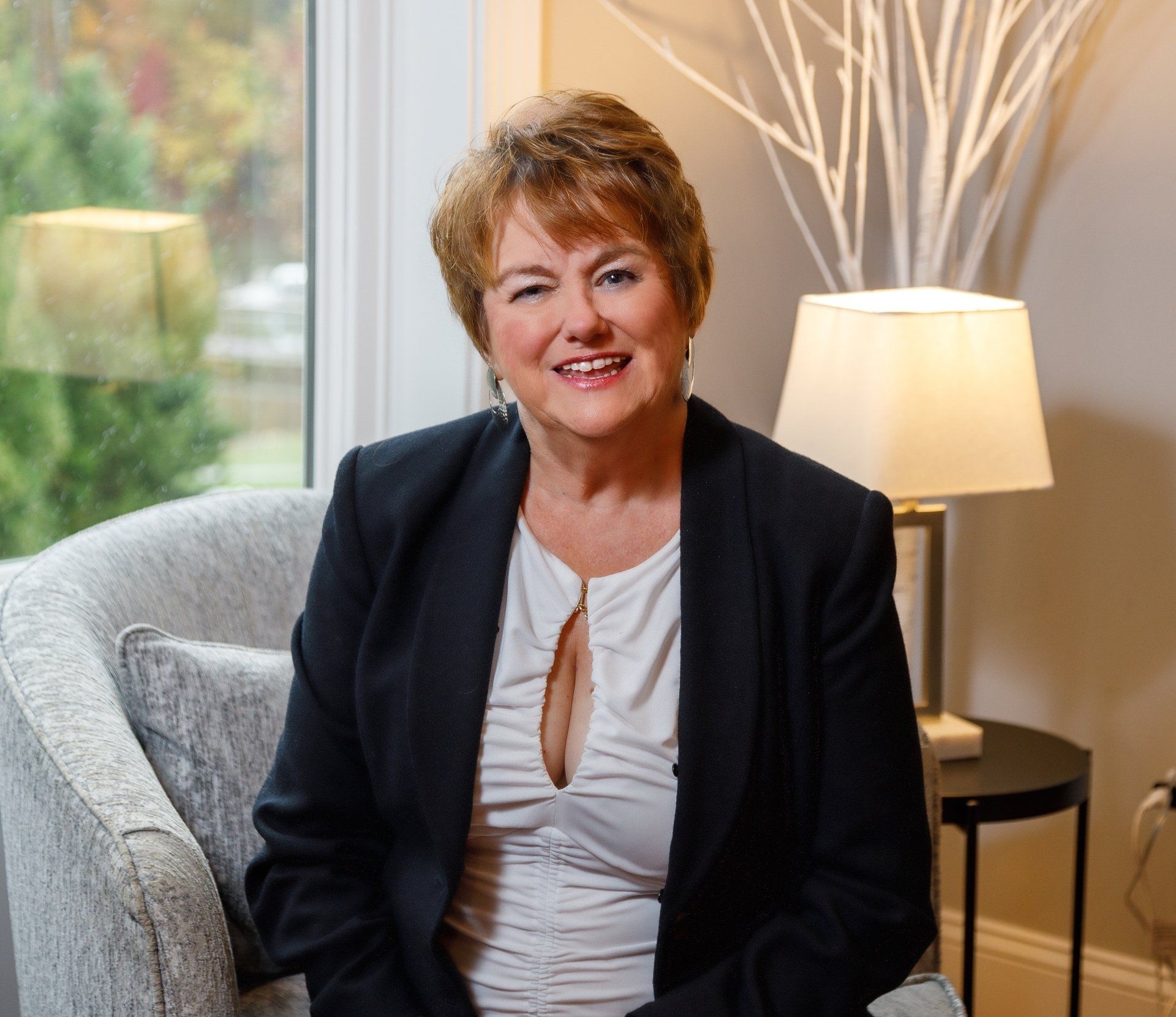 Woman in dark blazer sits in a chair near a window, smiling.