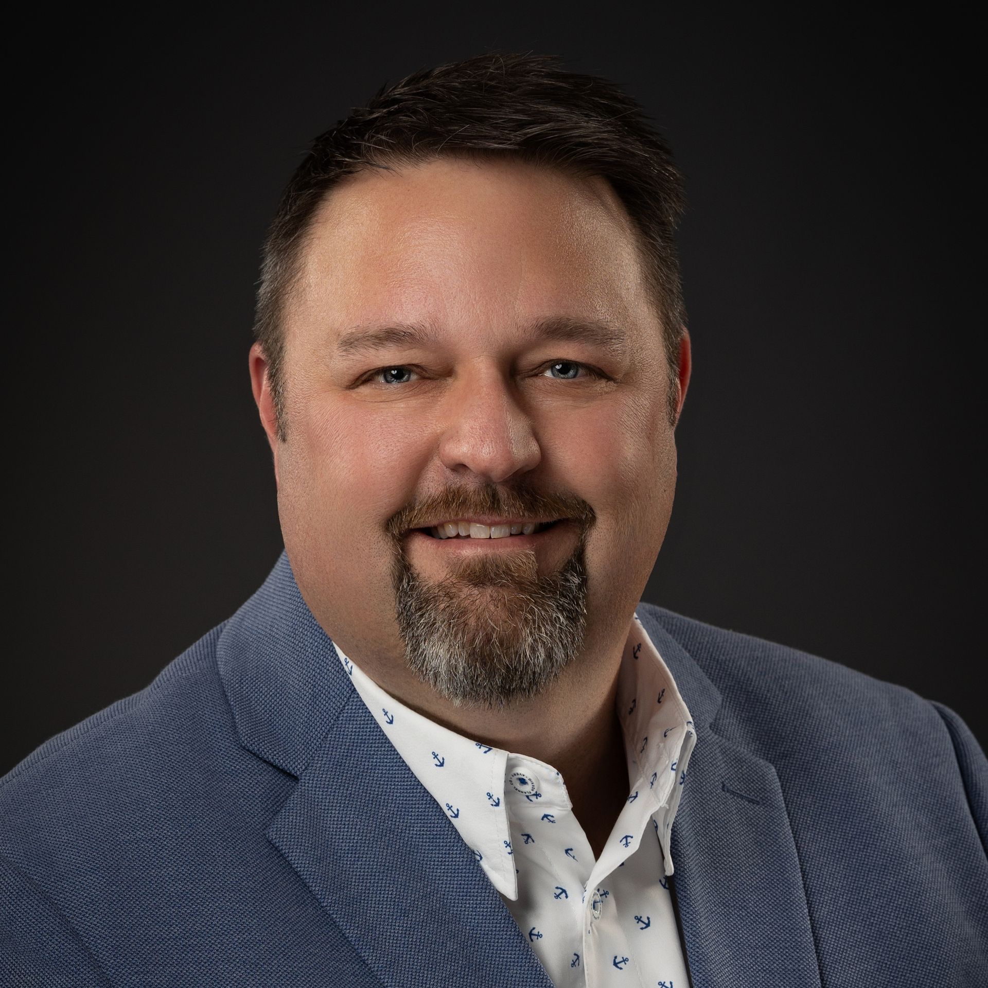 Man with beard smiles, wearing blue blazer and patterned white shirt; dark background.