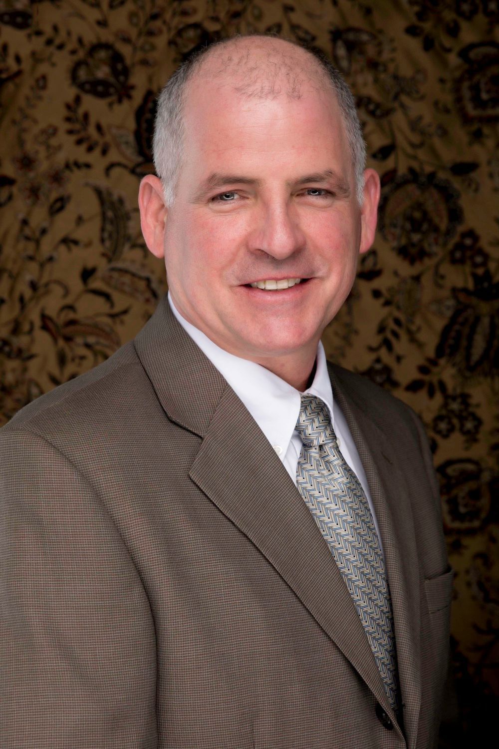 Man in a brown suit smiles at the camera, against a patterned background.