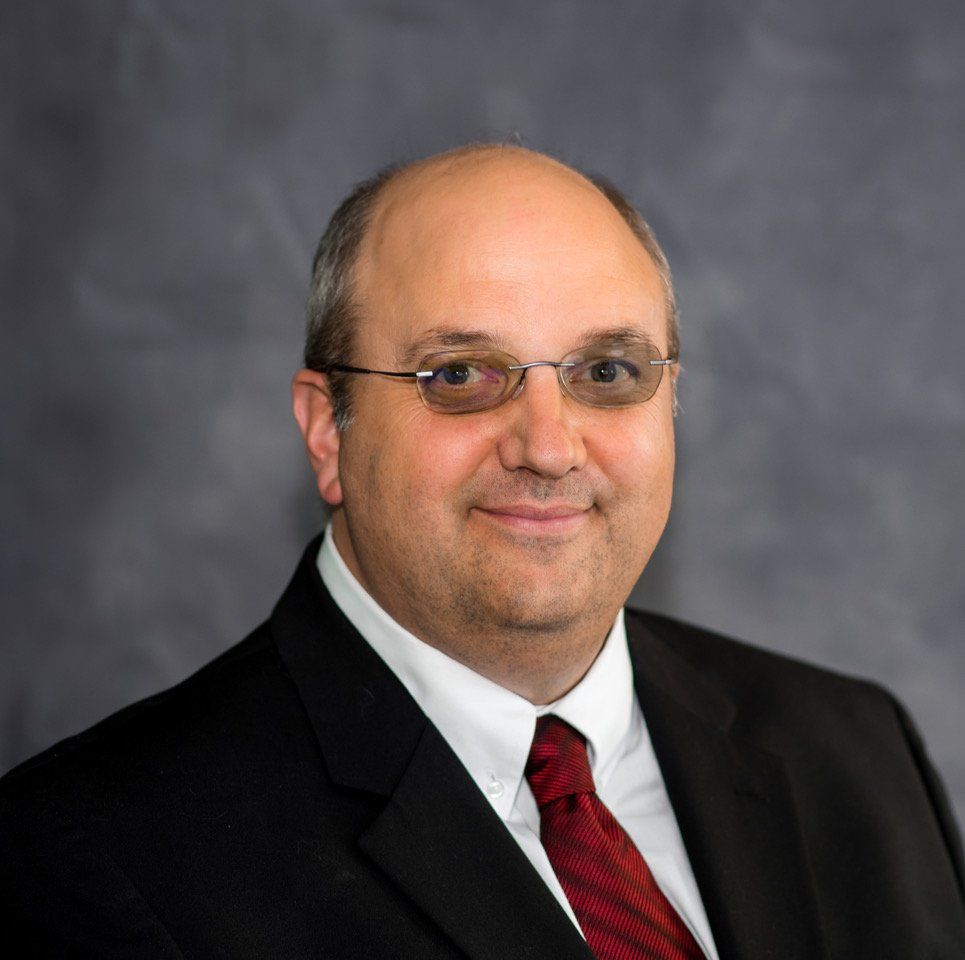 Middle-aged man with glasses in a suit and tie, smiling, against a gray background.