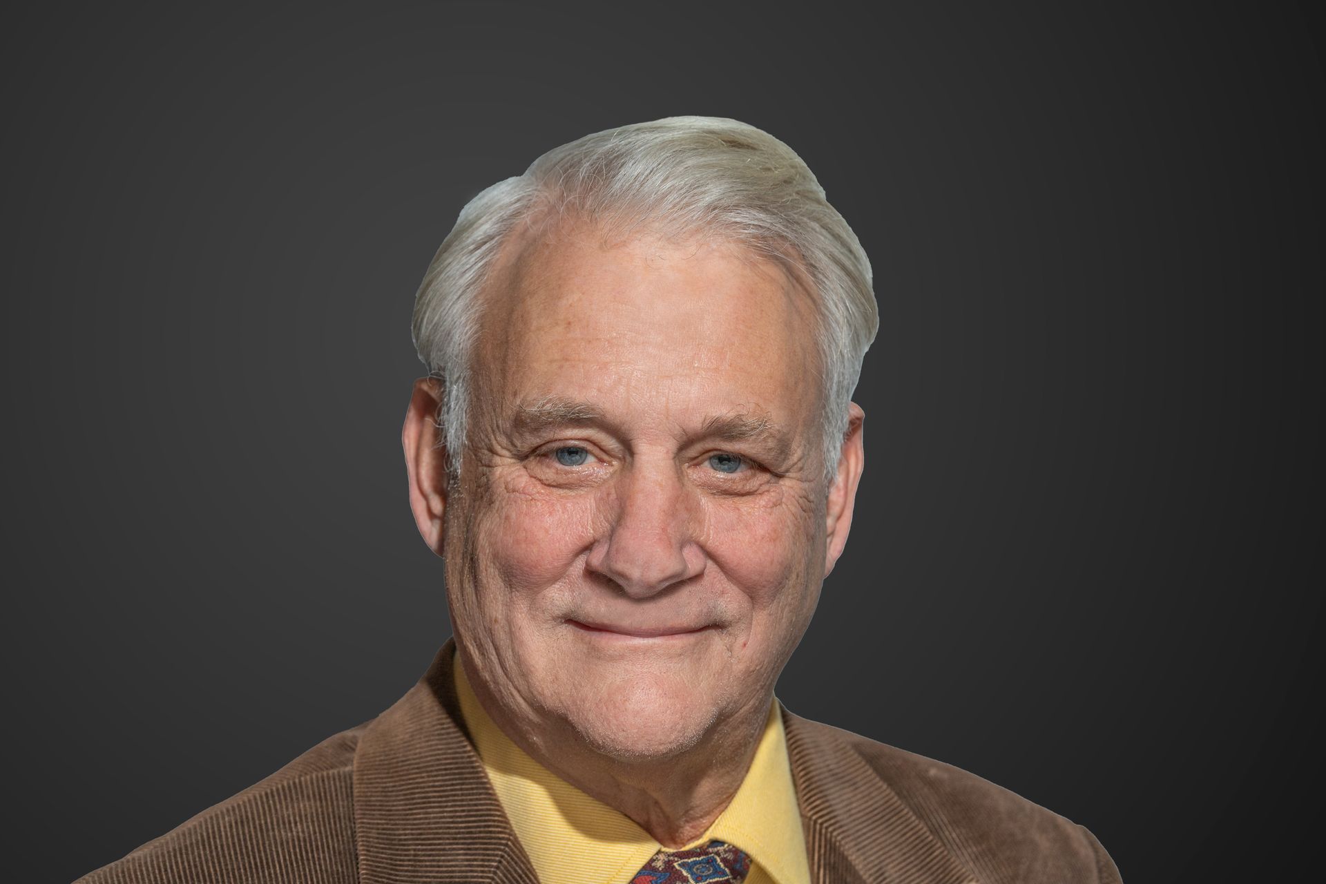 Man with gray hair and a brown corduroy jacket smiles, on a gray background.