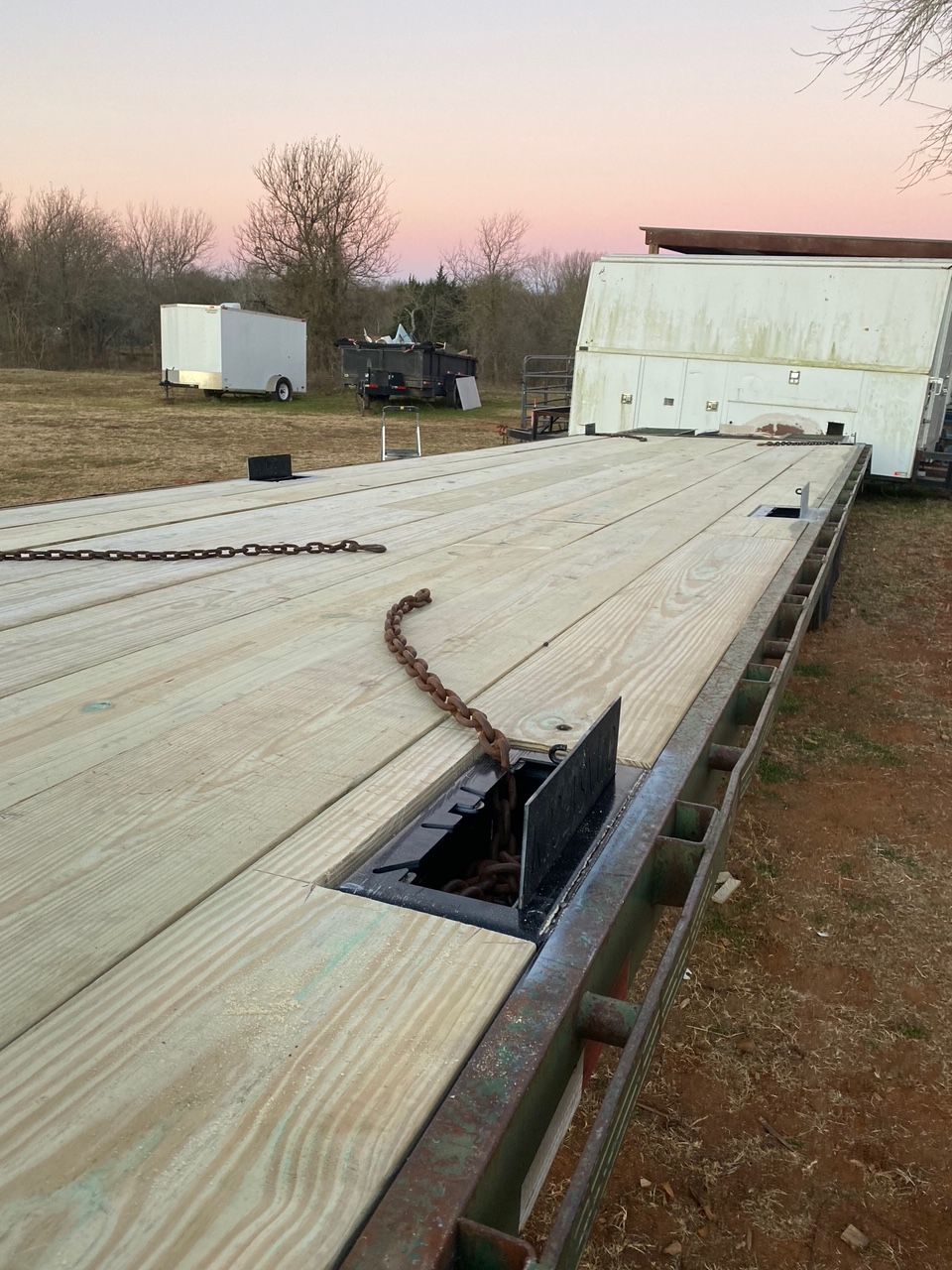 Flatbed trailer with new wood decking, chain, and storage hatch outdoors at dusk.