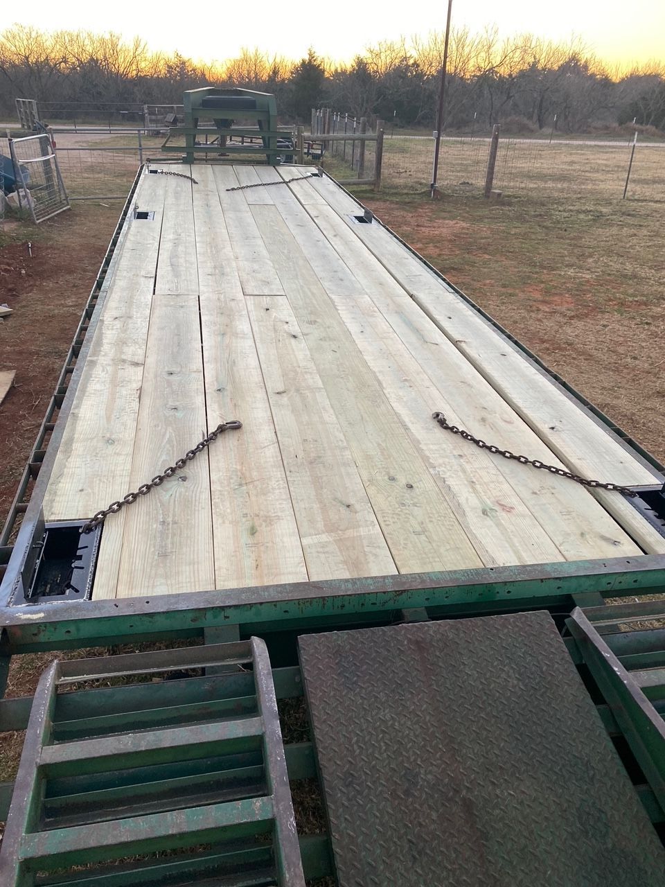 Flatbed trailer with wooden deck, chains, and ramp in a grassy field.